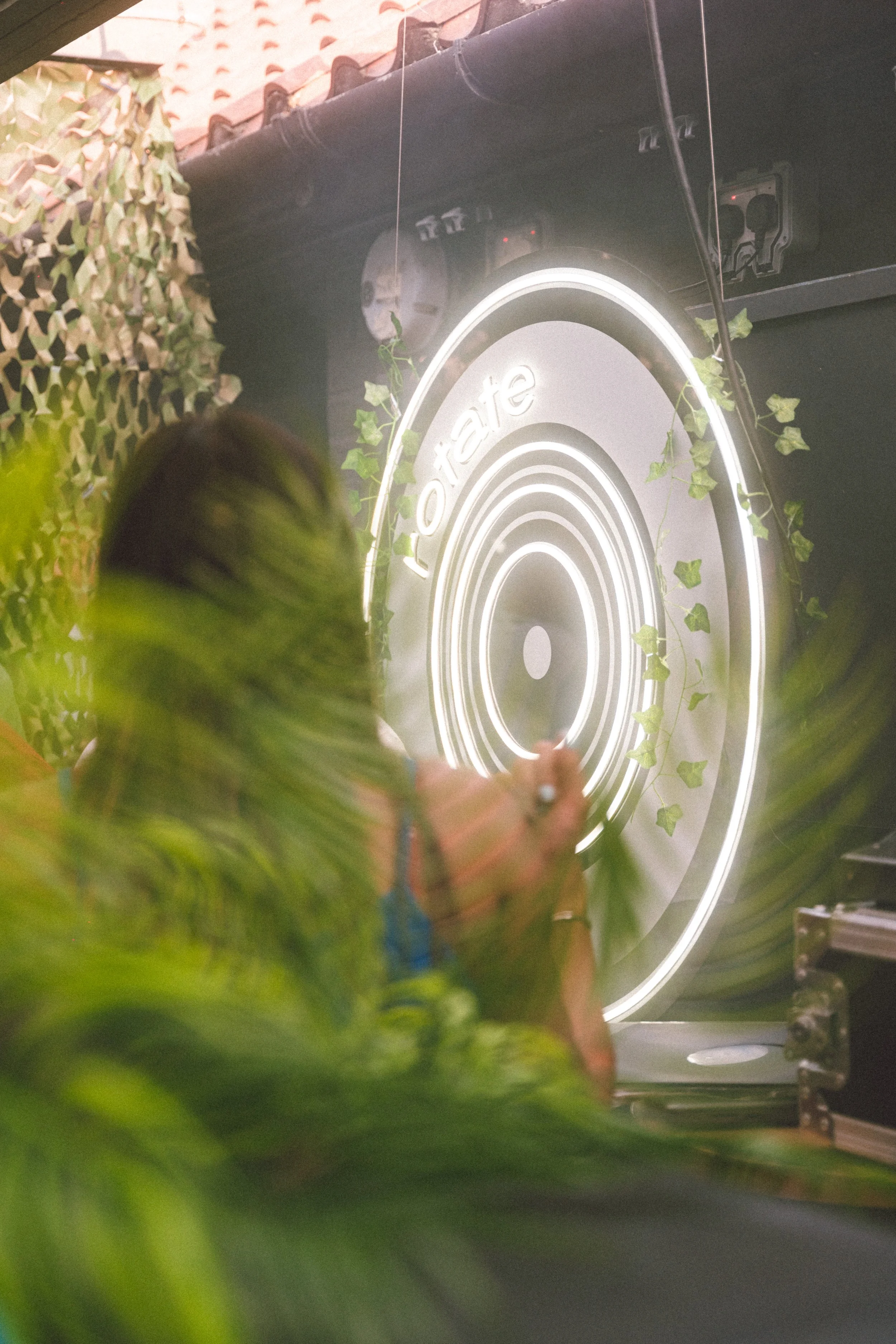 Person taking photo of illuminated logo on speaker, surrounded by greenery and textured wall, at an event with a modern, stylish setting.
