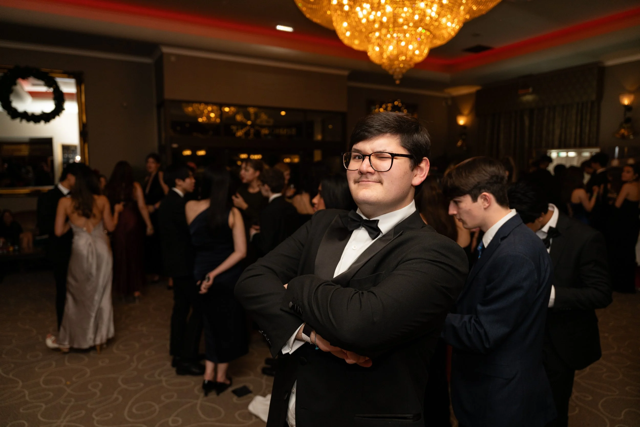 Young man in a tuxedo with glasses and a bow tie, standing with arms crossed and smirking, at a formal event with other people socializing in the background.