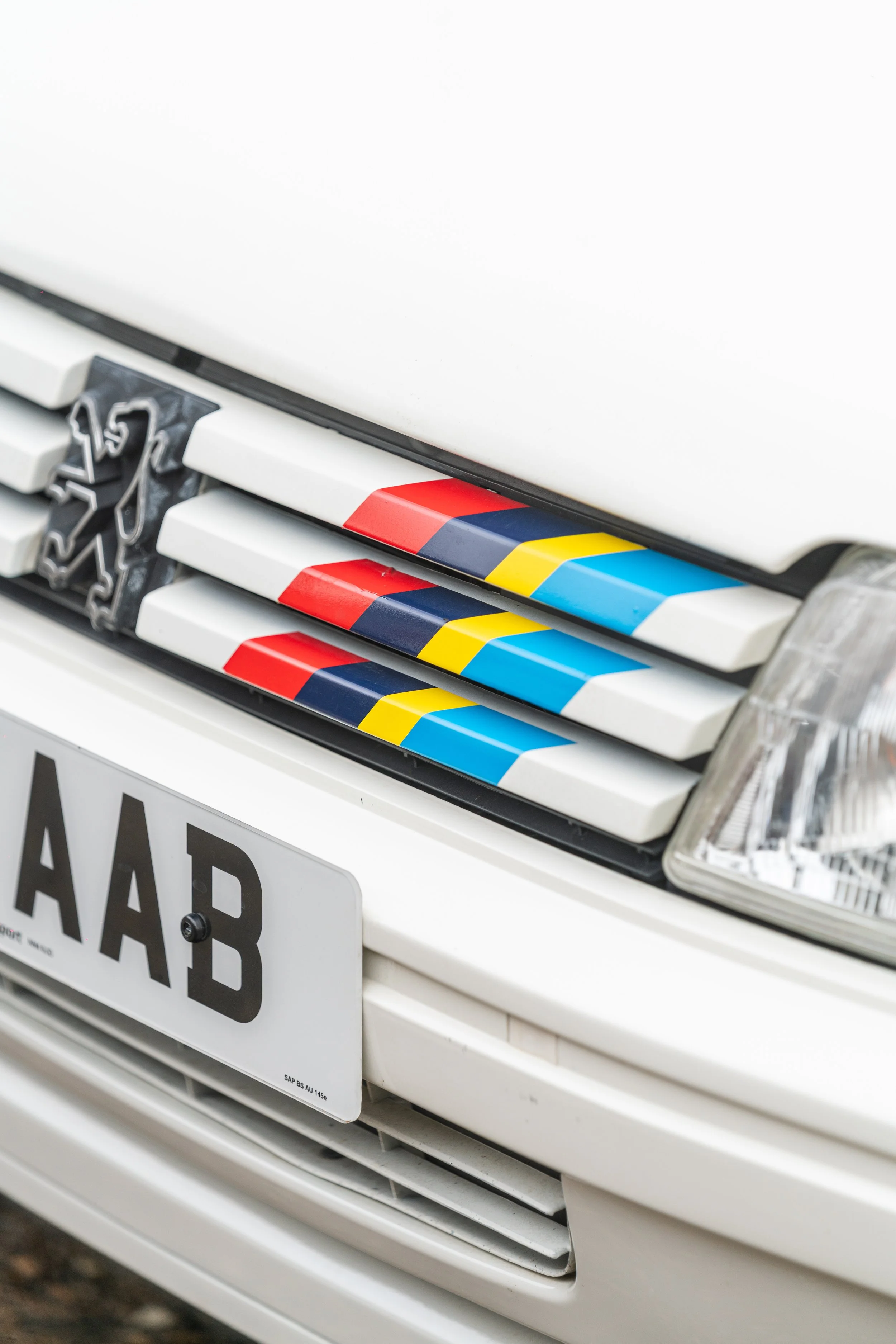 Close-up of the front grille and headlight of a white Peugeot car with colorful decals on the grille and a partially visible license plate reading 'AAB'.