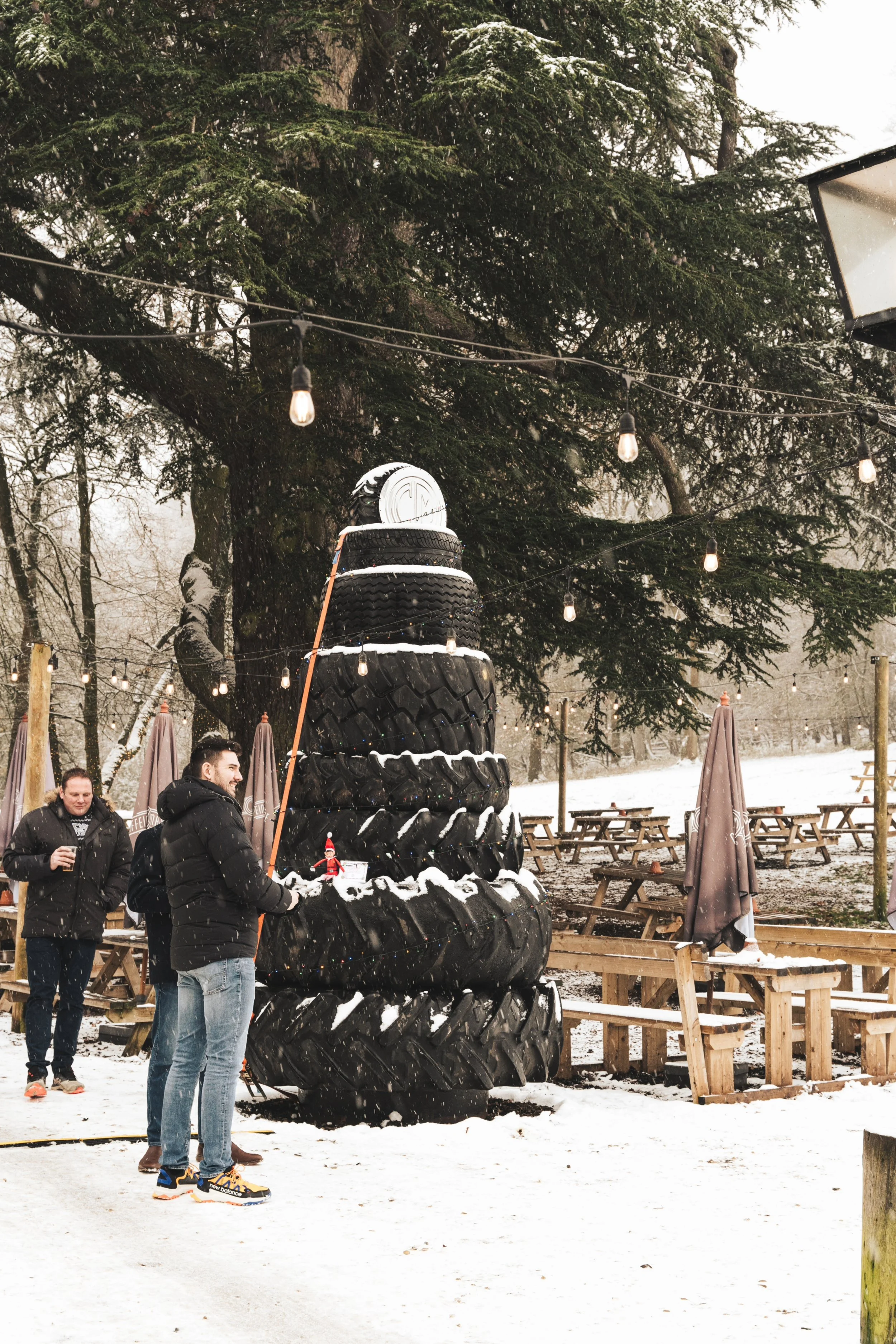 People standing near a sculpture made of stacked tires in a snowy outdoor setting with string lights and trees.