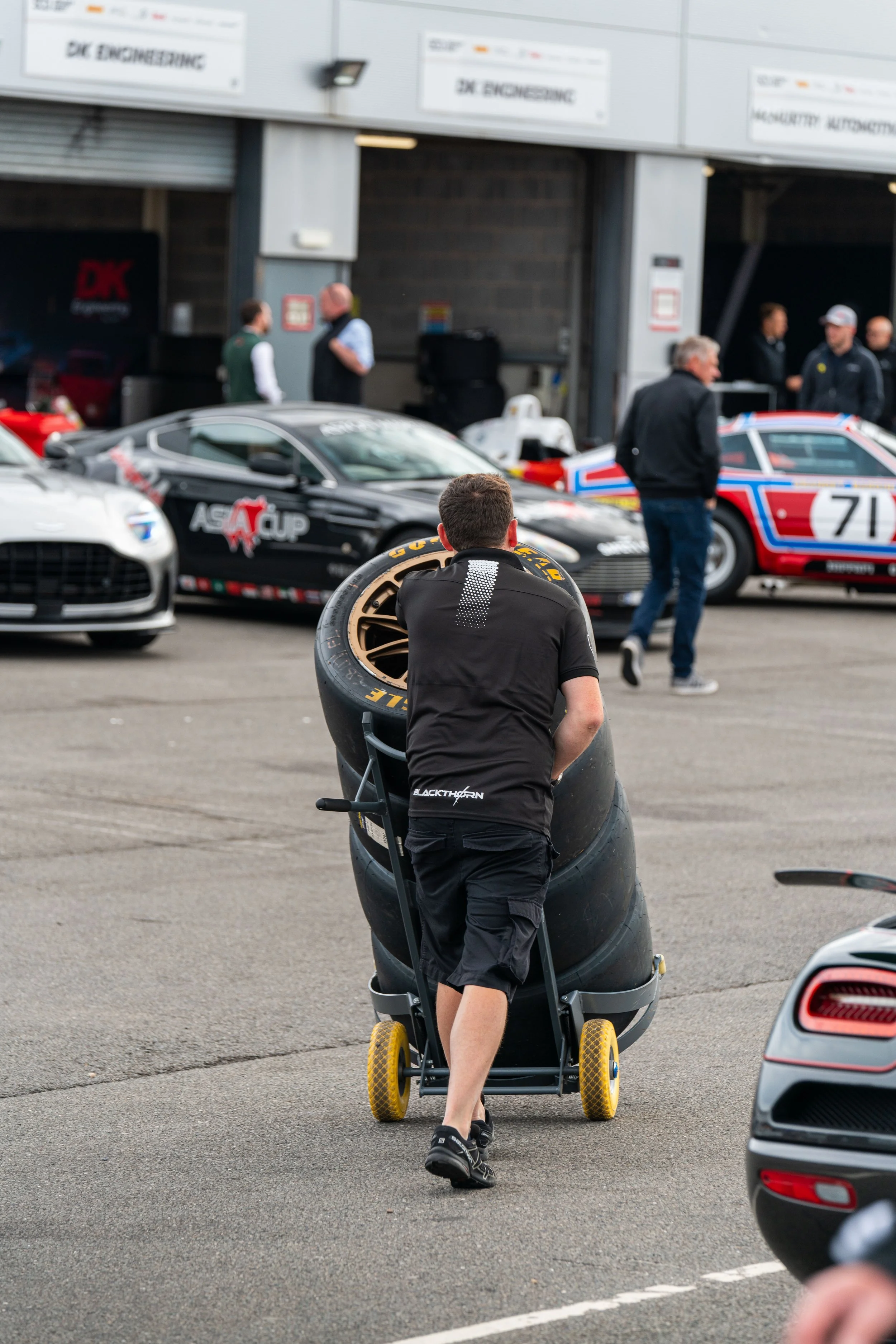 A person in black clothing pushing a tire cart with a stack of racing tires at a race car paddock, with race cars and people in the background.