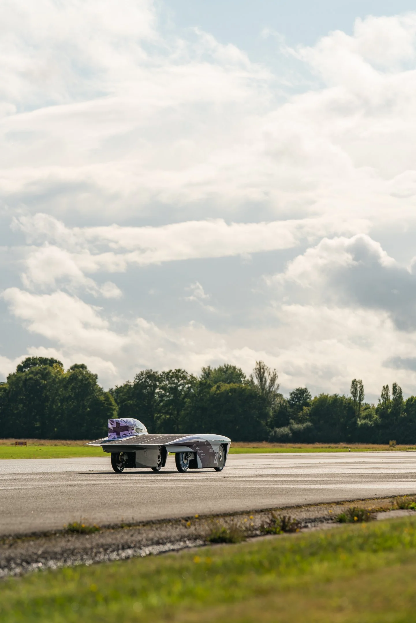 A futuristic car with a sleek black body and purple accents on an open roadway, with a background of green trees and a partly cloudy sky.