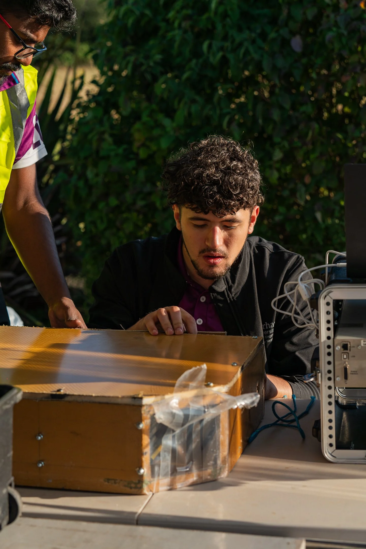 Two young men working outdoors with electronic equipment and a wooden box, one seated and examining, the other standing nearby, with green trees in the background.