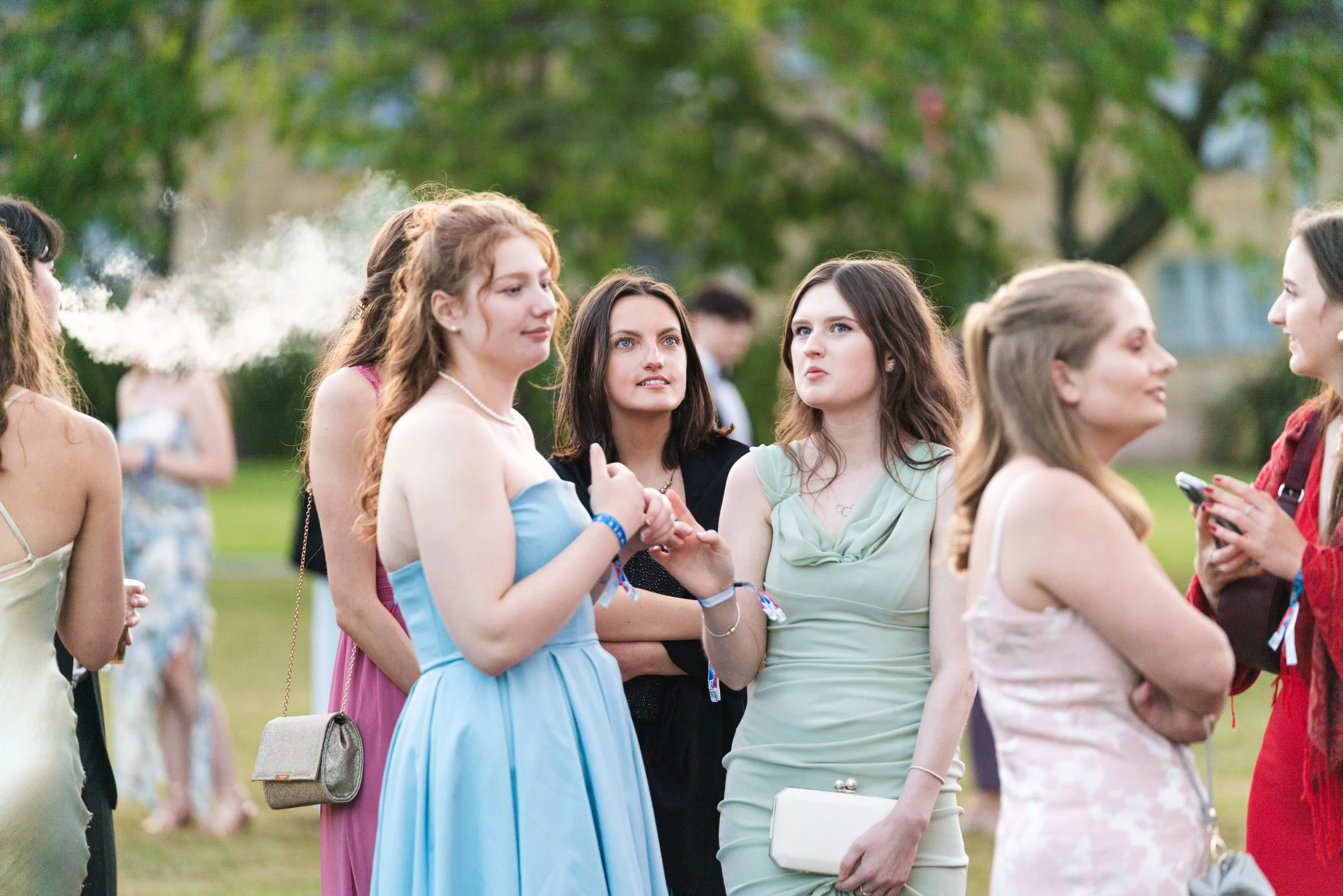 Group of young women dressed in formal and semi-formal attire socializing outdoors, with trees and grass in the background.