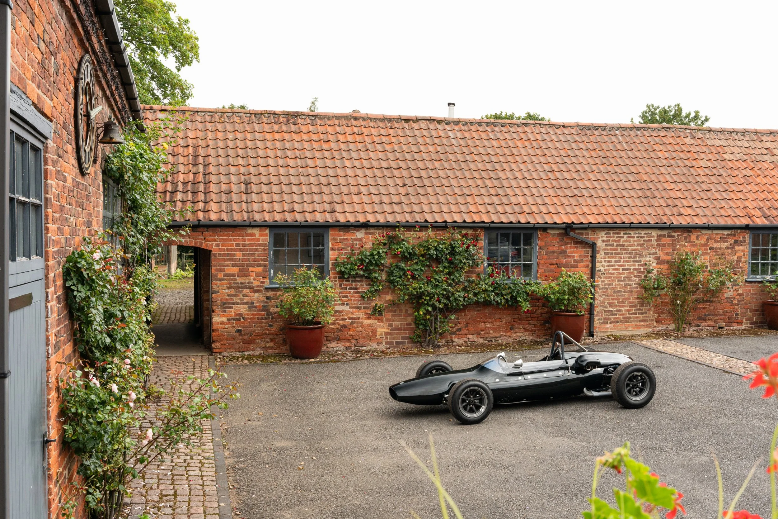 A vintage black race car parked in an outdoor courtyard with a brick building in the background. The building has a red tiled roof, windows with black frames, and potted plants on the ground.