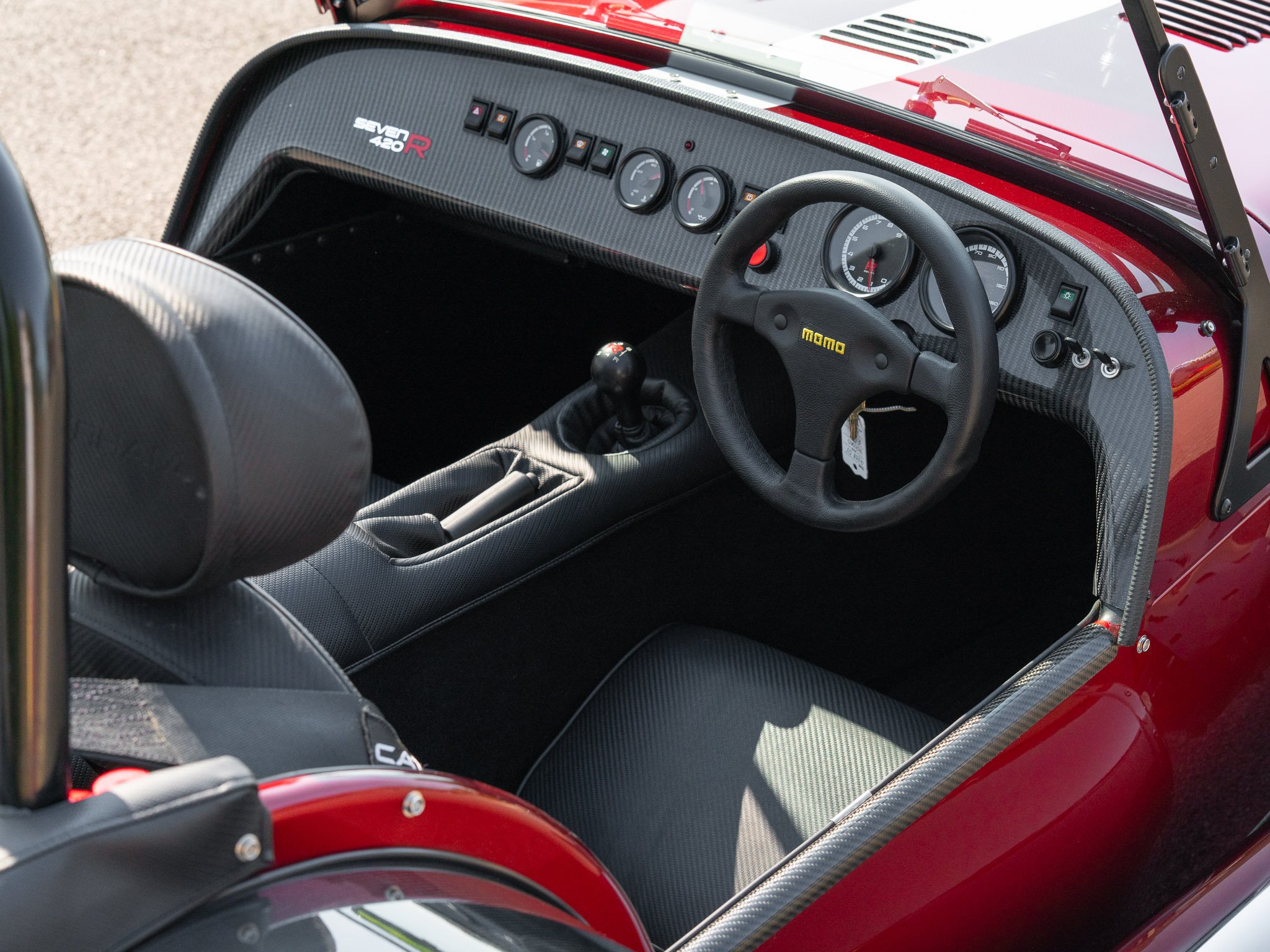 Interior of a race car with a carbon fiber dashboard, a racing seat, a MOMO steering wheel, and a gear shifter, with multiple gauges on the dashboard.