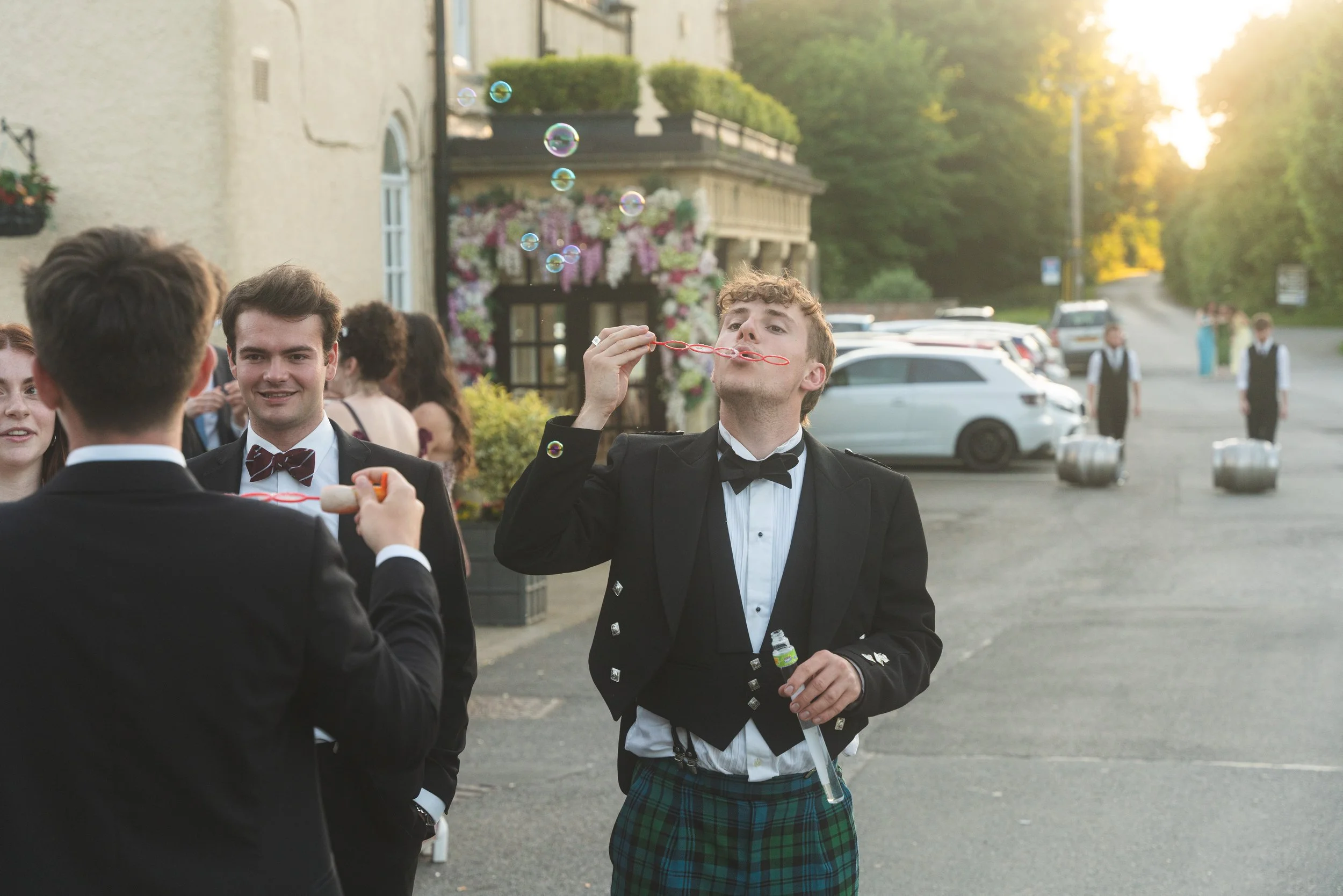 Group of people dressed in formal attire, with one person blowing bubbles outdoors during evening.