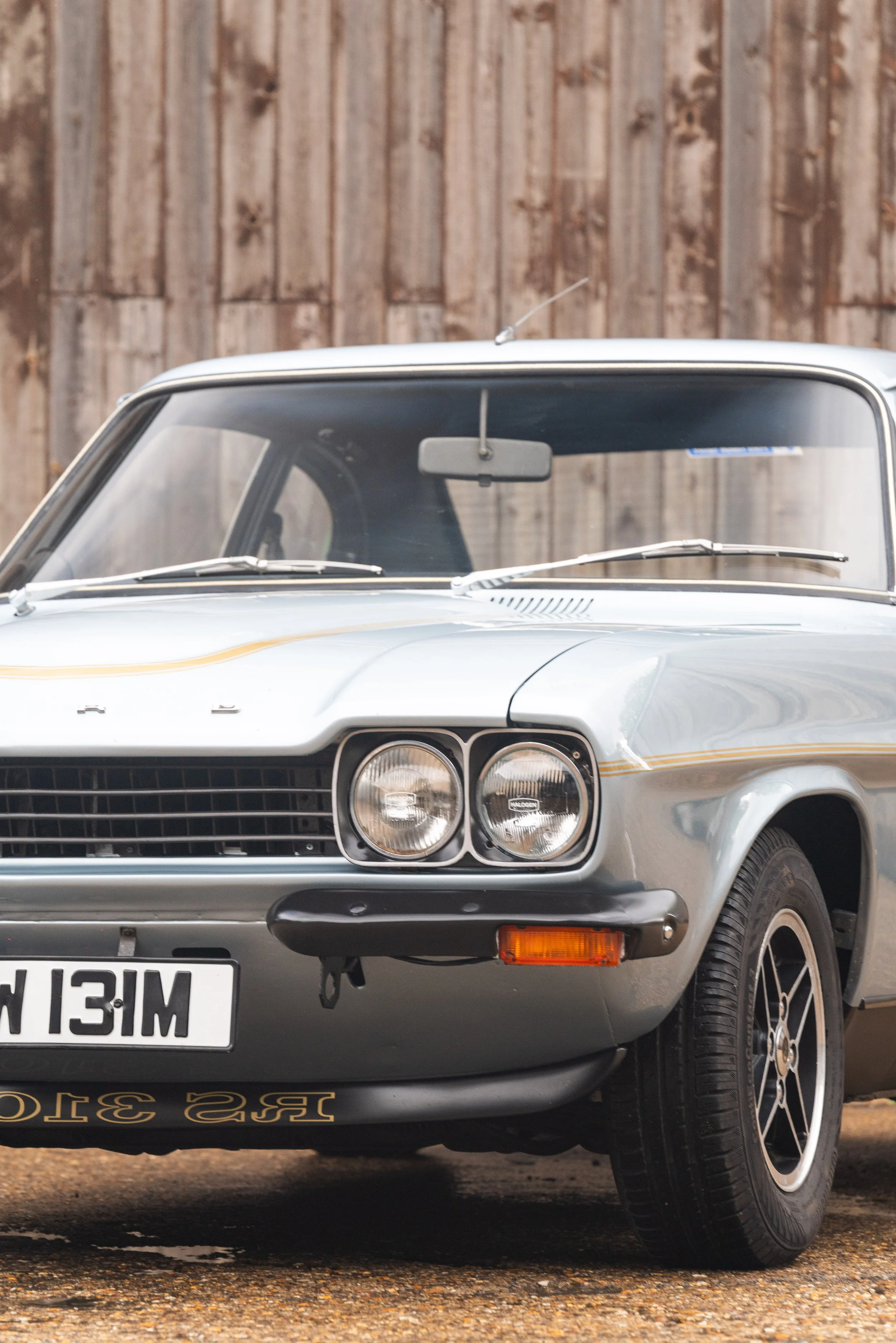 Front view of a vintage silver classic car parked outdoors with a wooden fence in the background.