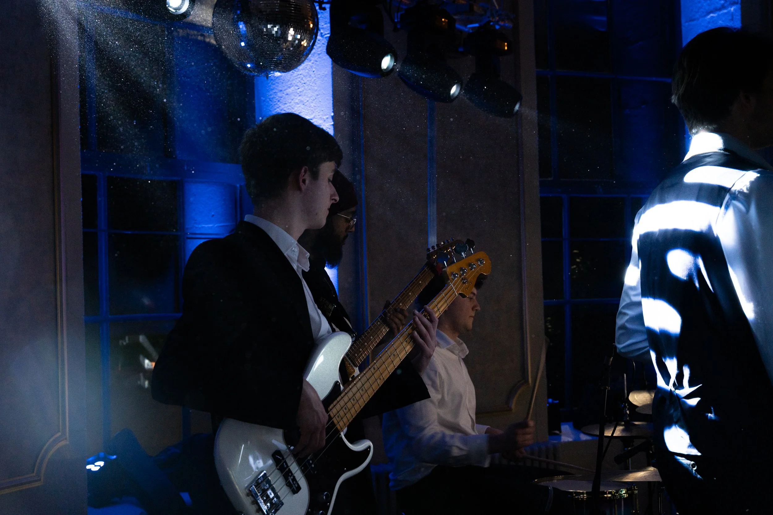 Musicians performing at a live event, with one playing bass guitar, another on drums, and a third person in the foreground, lit by blue stage lighting.