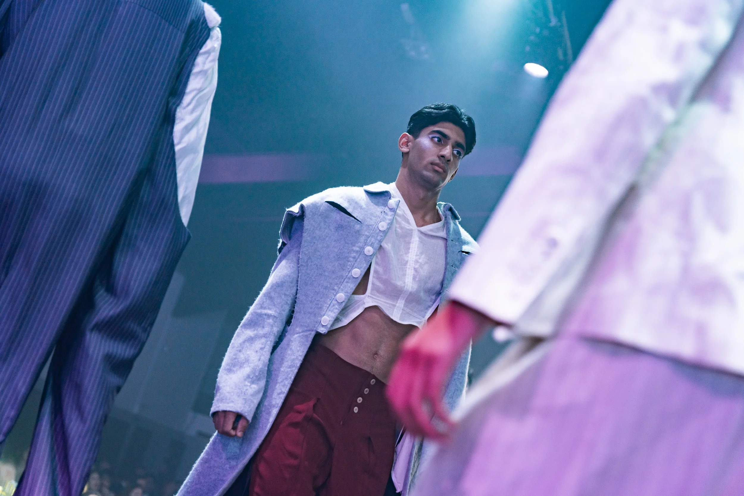 A man with dark hair and a serious expression standing among others, wearing a long coat, a sheer top revealing his midriff, and maroon pants in a dimly lit indoor space.