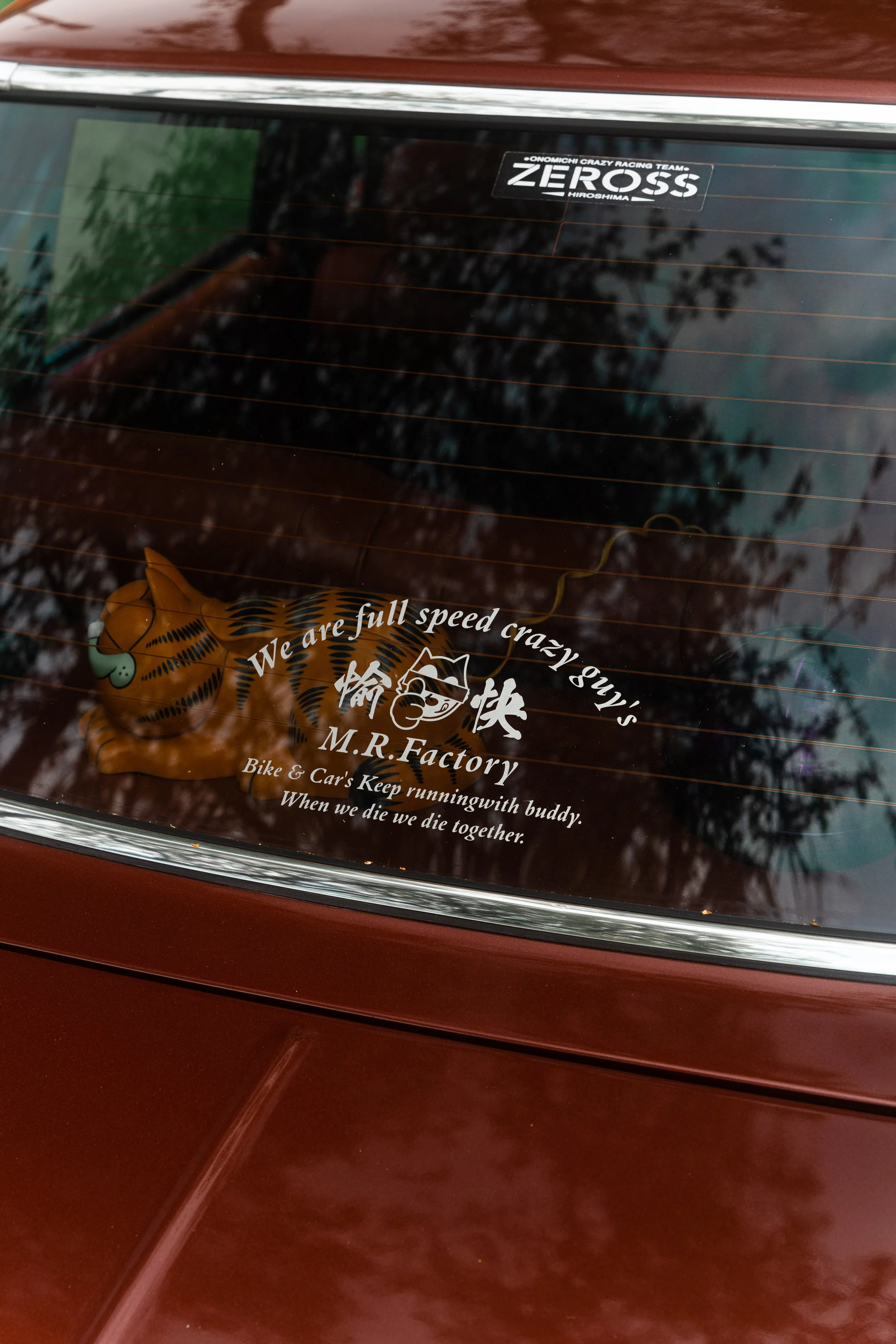Back window of a car with multiple stickers, including one with a tiger cartoon and text about speed and camaraderie, and another sticker with the word 'ZEROSS' near the top.