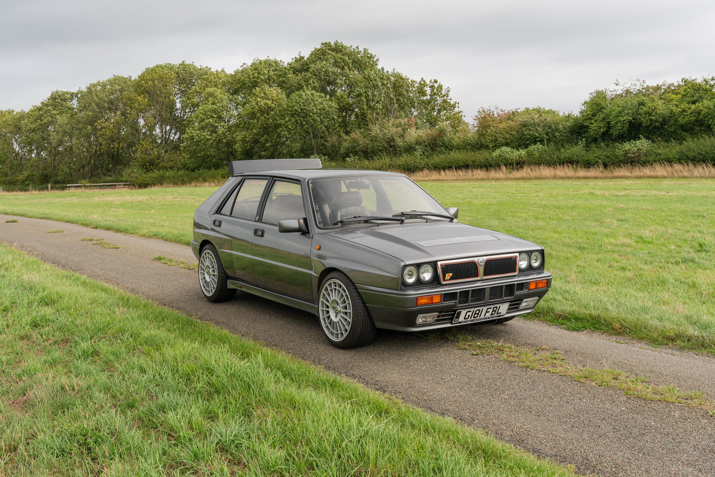 A classic gray hatchback car parked on a narrow paved road beside green grass and trees under a cloudy sky.