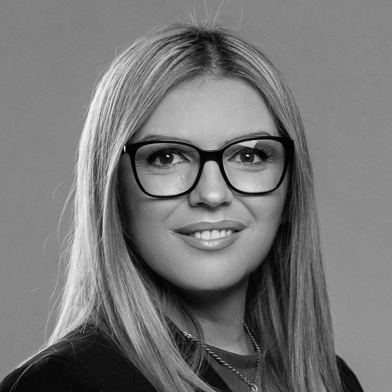 Black and white photo of a smiling woman with glasses and earrings, wearing layered necklaces, indoors with blurred background.