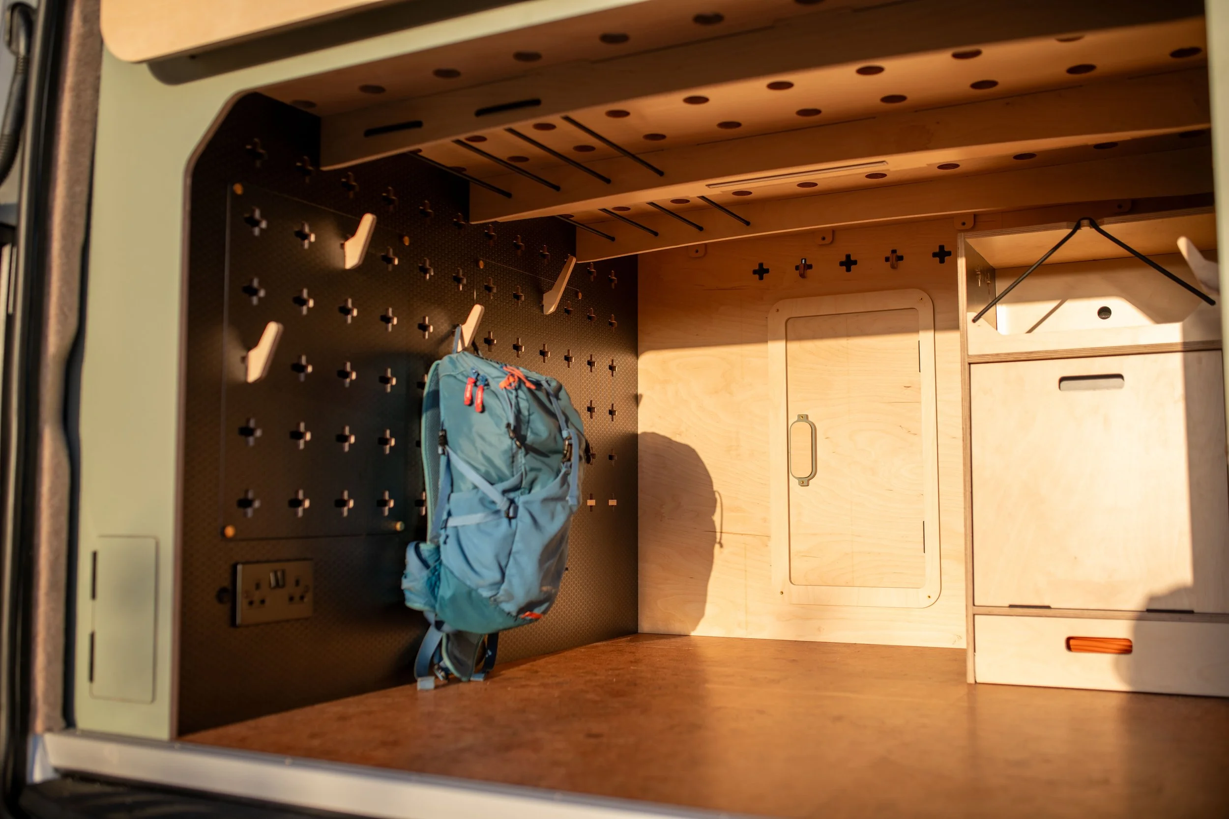 Empty wooden storage compartment with pegboard and hooks, a backpack hanging, and a small door in the back wall.