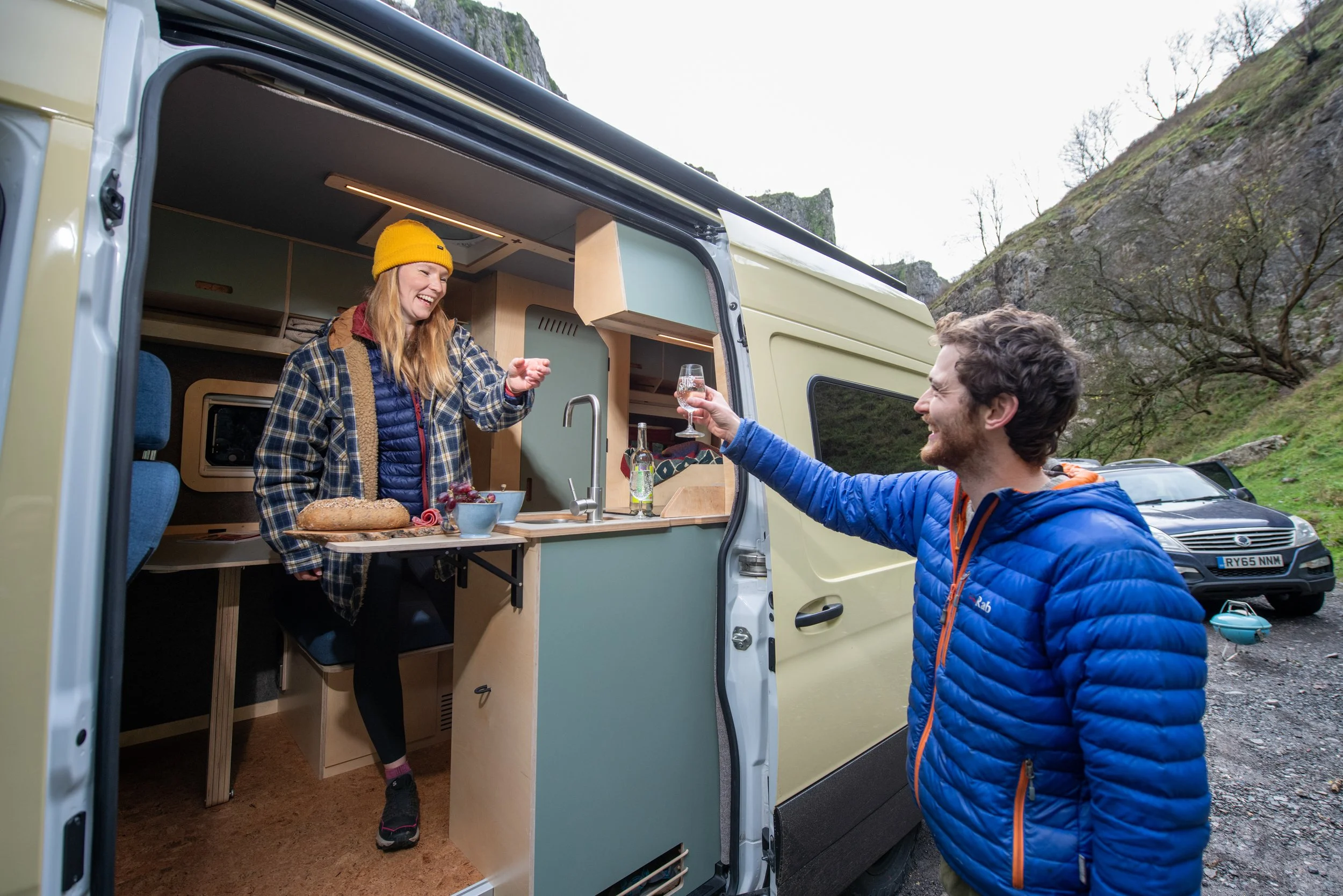 Two friends sharing drinks at the entrance to the kitchen of a Fern campervan, using the slide-out table to serve food and host at a campsite.