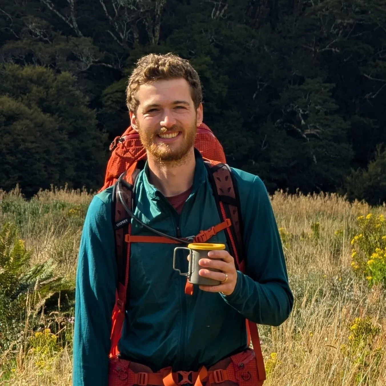 A man with a beard smiling outdoors in a grassy field, holding a camping mug, wearing a blue jacket and a large red backpack.