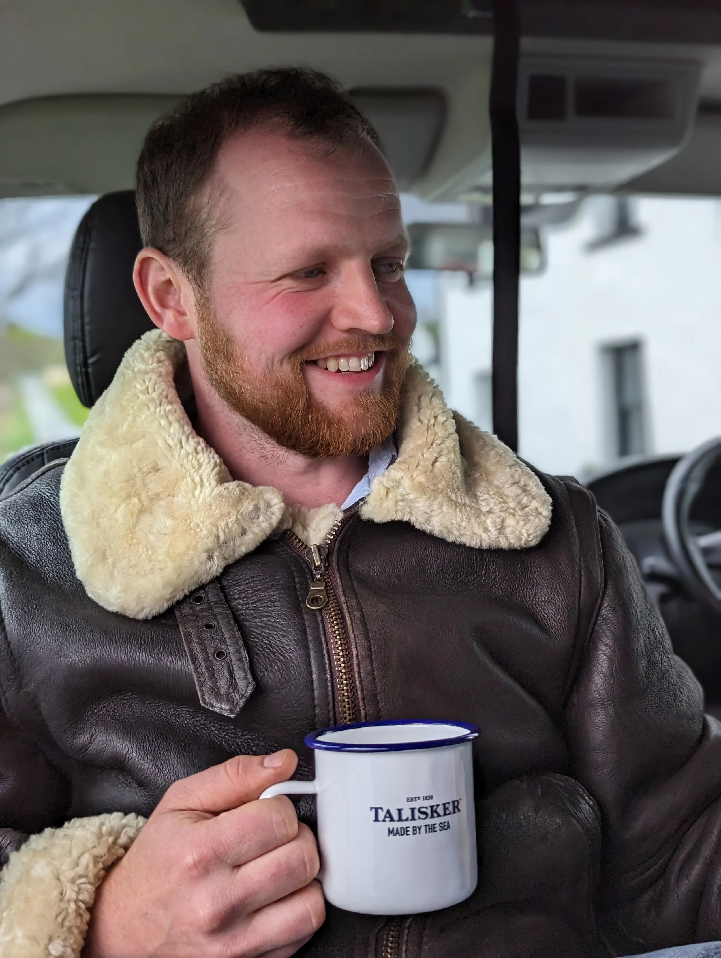 A man with a beard and red hair smiling, sitting in a vehicle, holding a mug that reads 'TALISKER MADE BY THE SEA'.