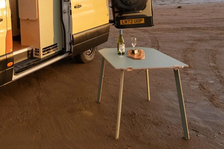 Freestanding Fern campervan table set up on the beach with bread and wine, creating an easy outdoor dining space beside the van.