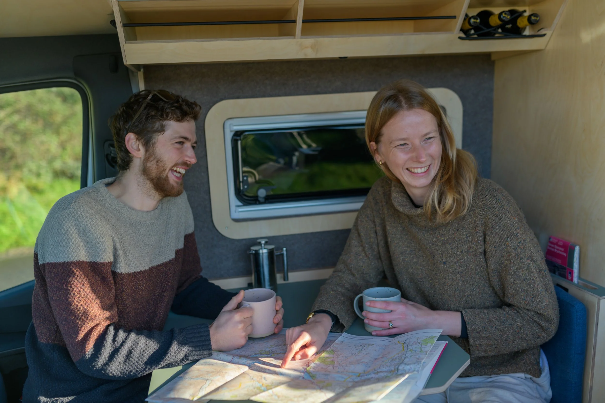 A couple in the seating area of a Fern campervan, with mugs and a paper map, planning their next route while in comfort indoors.