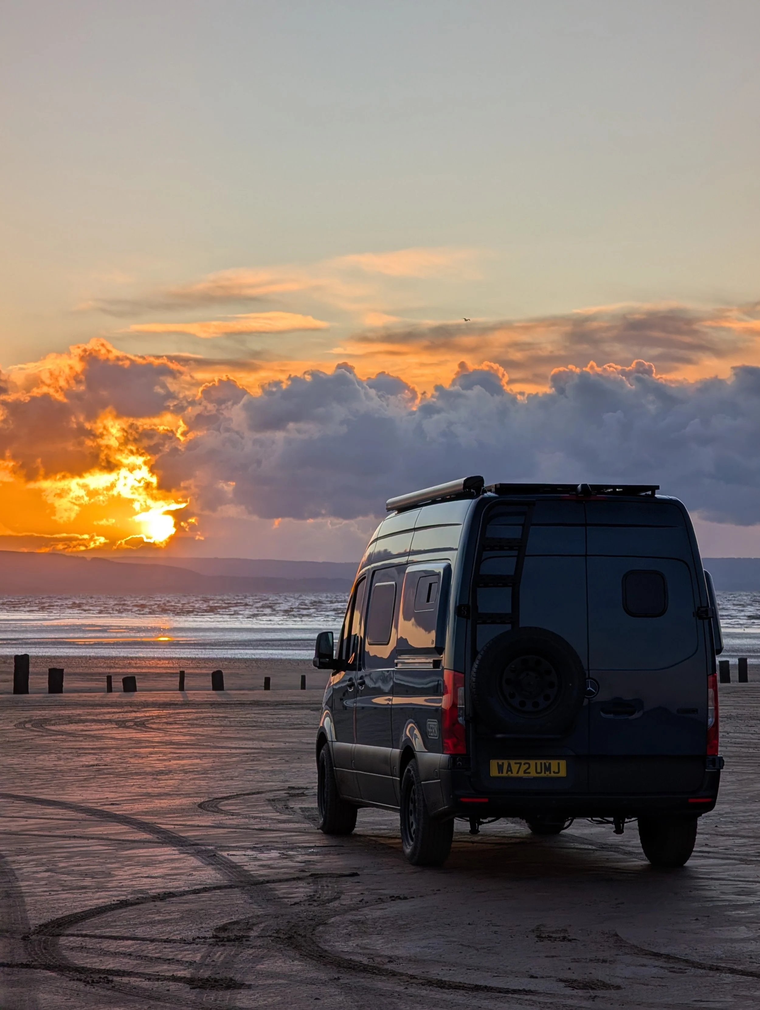 A van parked on a sandy beach during sunset with the ocean and cloudy sky in the background.