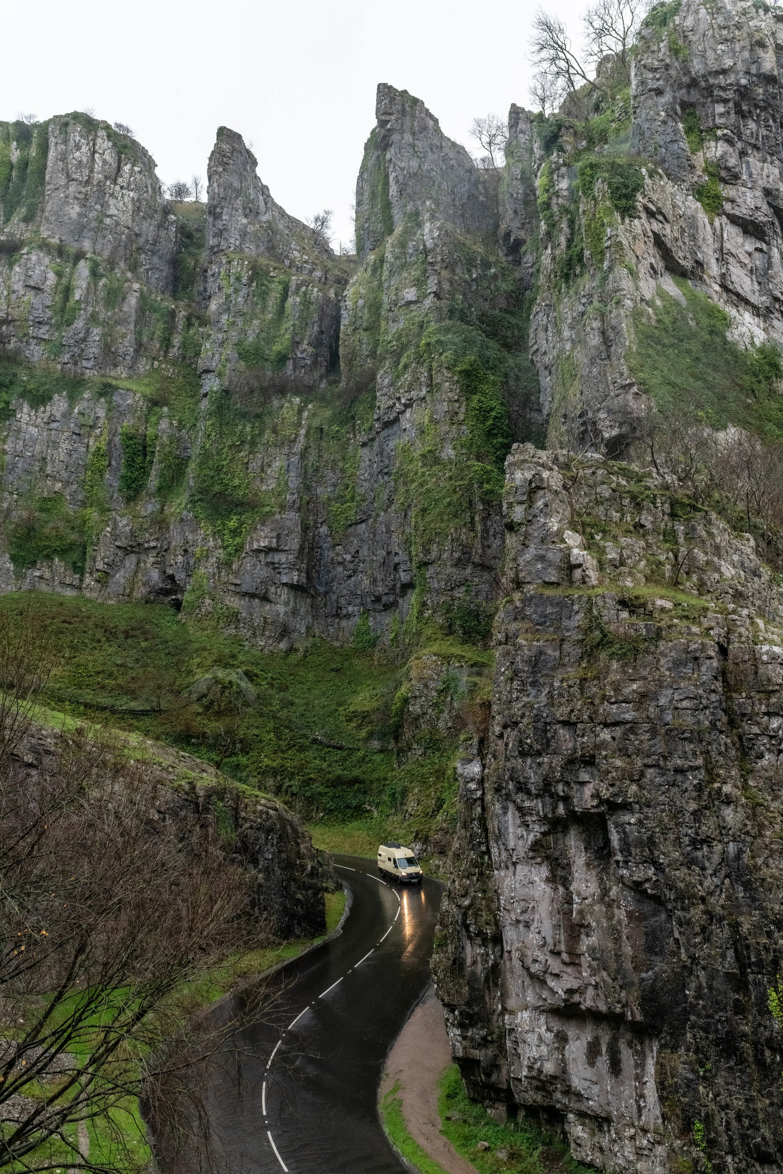 A white campervan is driving along a winding route, in the autumn rain. The road leads through Cheddar Gorge, a rocky canyon with steep cliffs in Somerset.