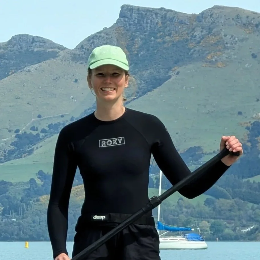 A woman smiling while holding a paddle near a lake with sailboats and mountains in the background.