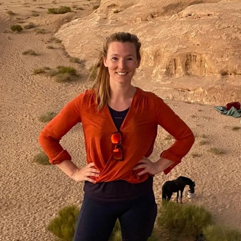 Lucy, Fern Campervans' Director of Assets, is standing outdoors in a Jordanian desert with rocky cliffs, and horses grazing nearby. She is smiling and poses with hands on hips.