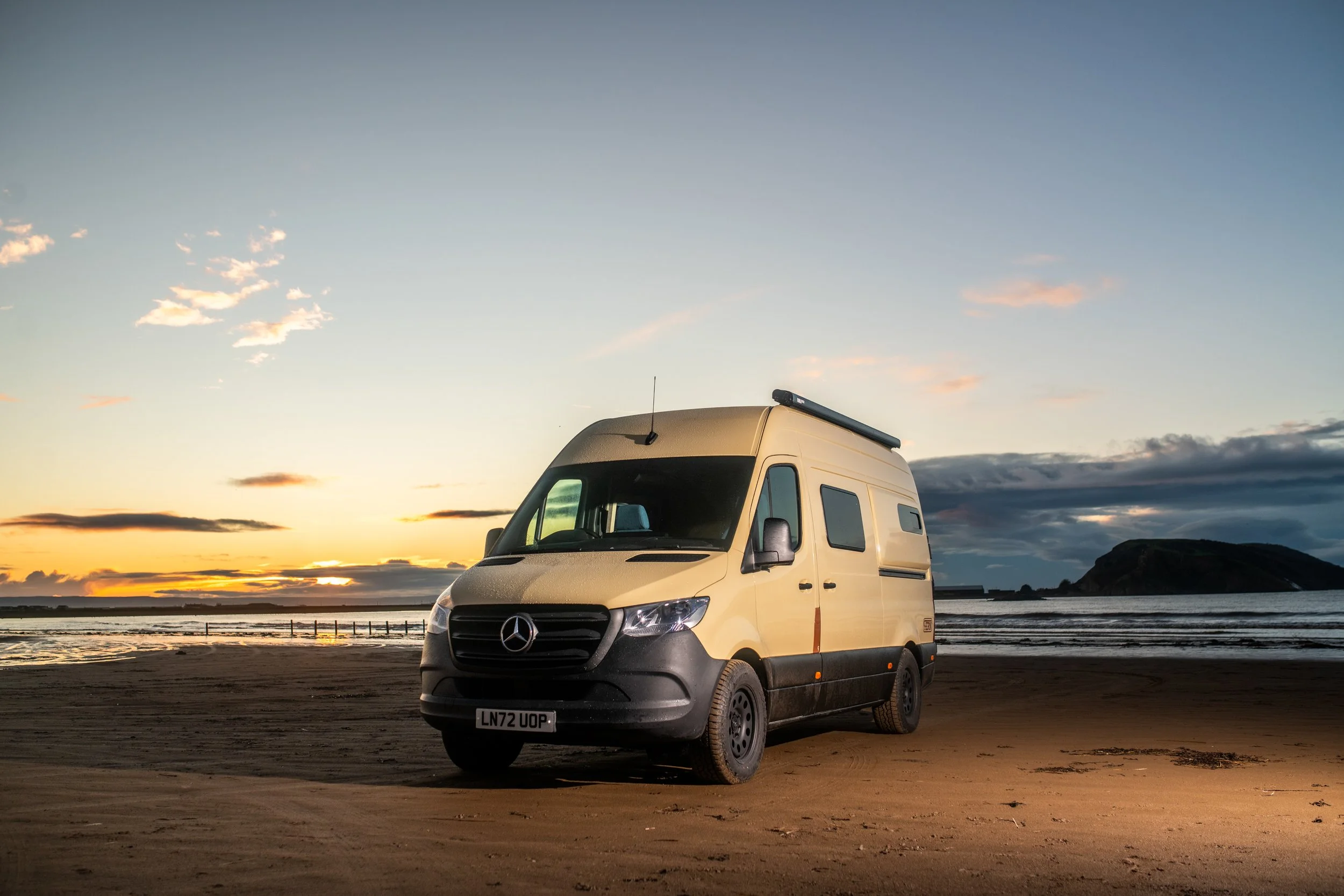 A beige Mercedes-Benz Sprinter van parked on a sandy beach during sunset with a hill in the background.