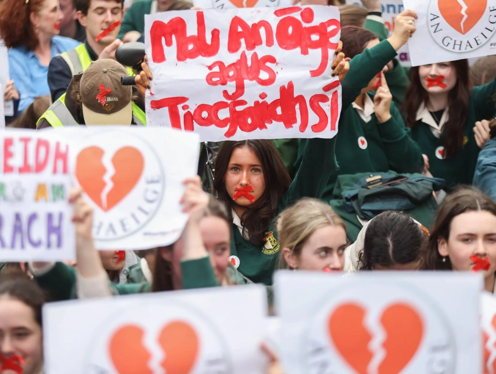 Group of young people at a protest or rally, some holding signs with messages and symbols, including a broken heart and Irish phrases, with some having red tape over their mouths.