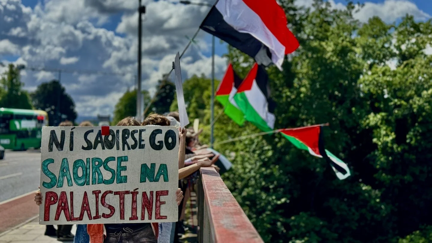Protesters holding a sign that reads 'Ni saoire go saorise na Palestina' during a demonstration with Palestinian flags in the background.