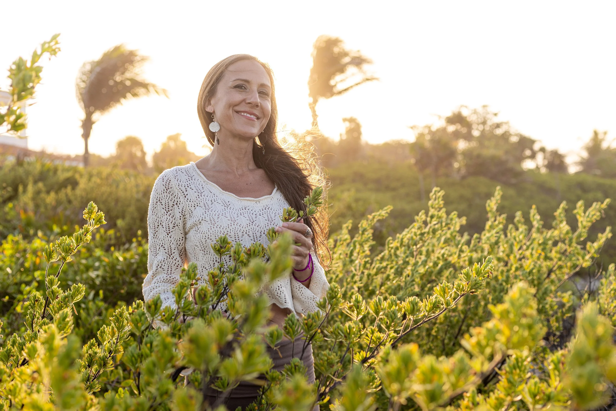 Jaime Fleres, wearing a cream-colored crochet top and earrings, stands among green shrubs outdoors during sunset.