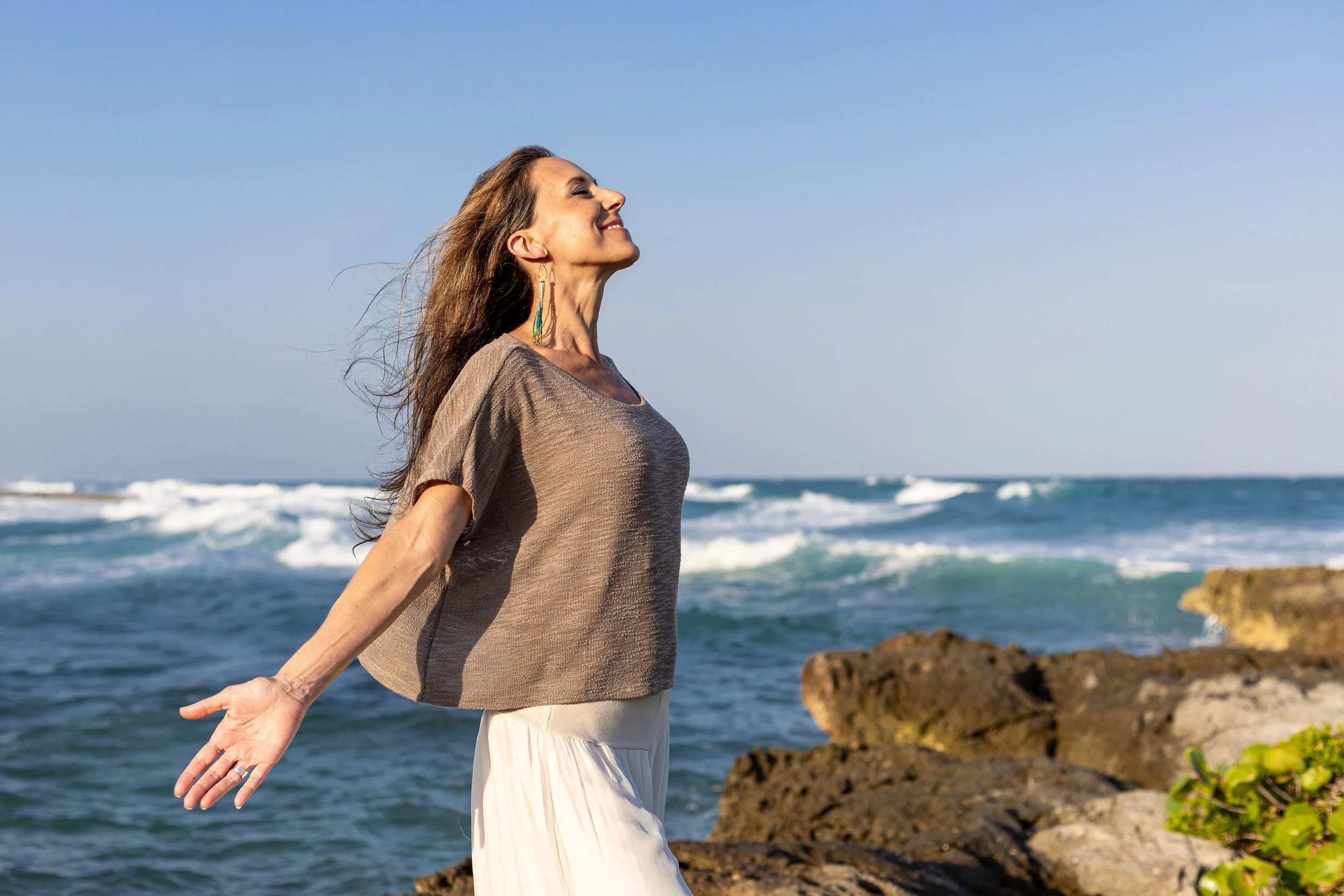 Jaime Fleres standing on rocks at the beach with her arms extended and face tilted upwards, smiling with eyes closed, ocean waves in the background, clear sky.