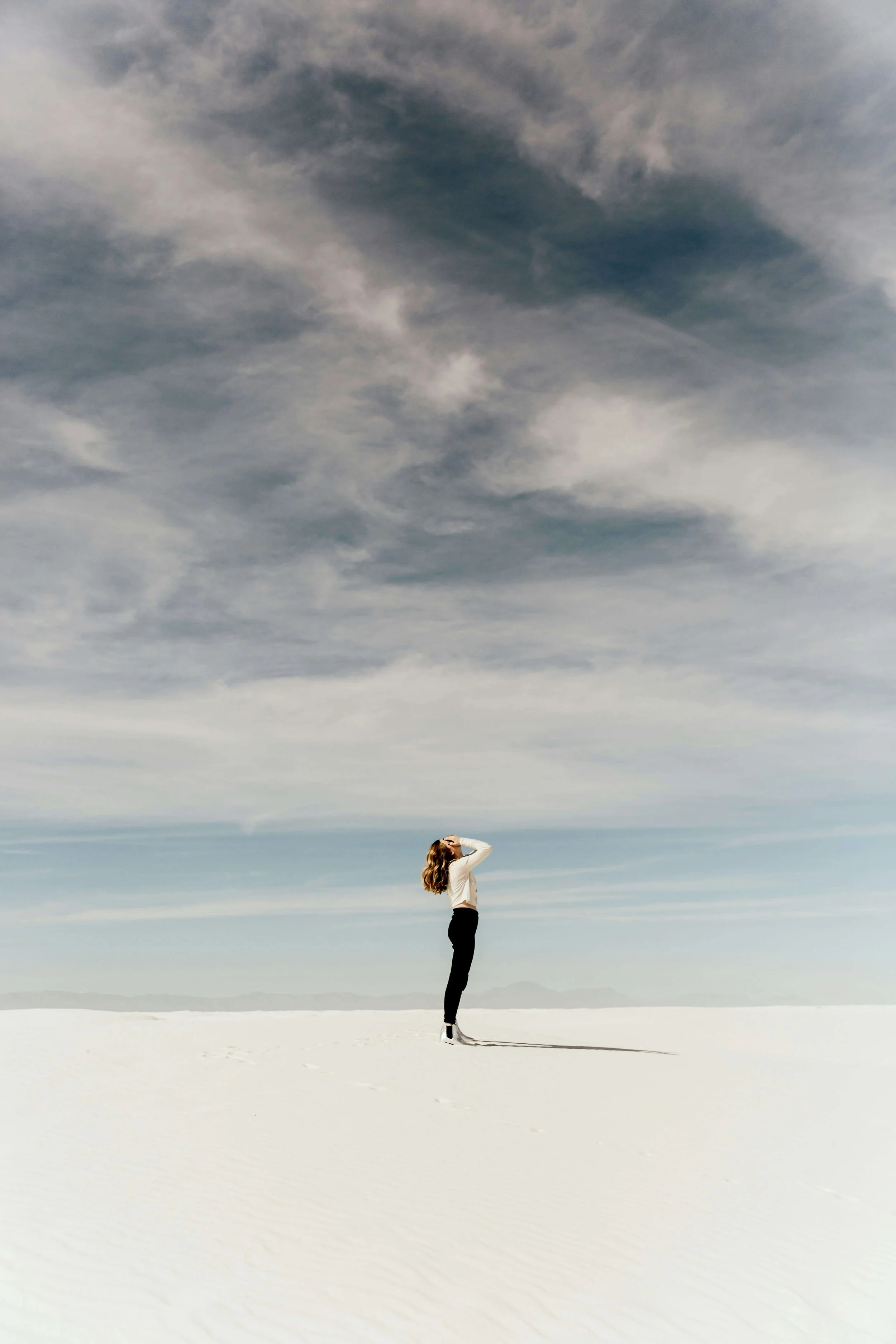 A woman standing alone on a vast, flat, white landscape under a cloudy sky, with her hand on her forehead as she looks into the distance.