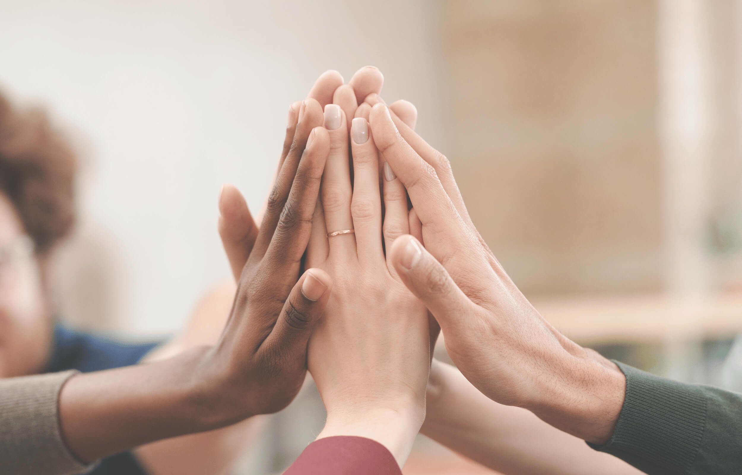 Multiple hands of diverse skin tones are stacked together in a gesture of unity or teamwork.