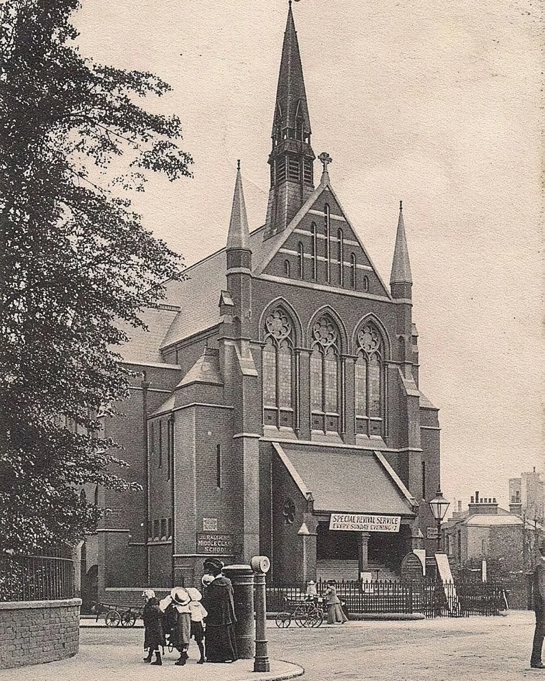 Historical black-and-white photo of a Gothic-style church with tall spire, large stained glass windows, and pointed arches. Pedestrians, including a woman with children and a man, are walking nearby, with bicycles and a lamp post visible in the scene.