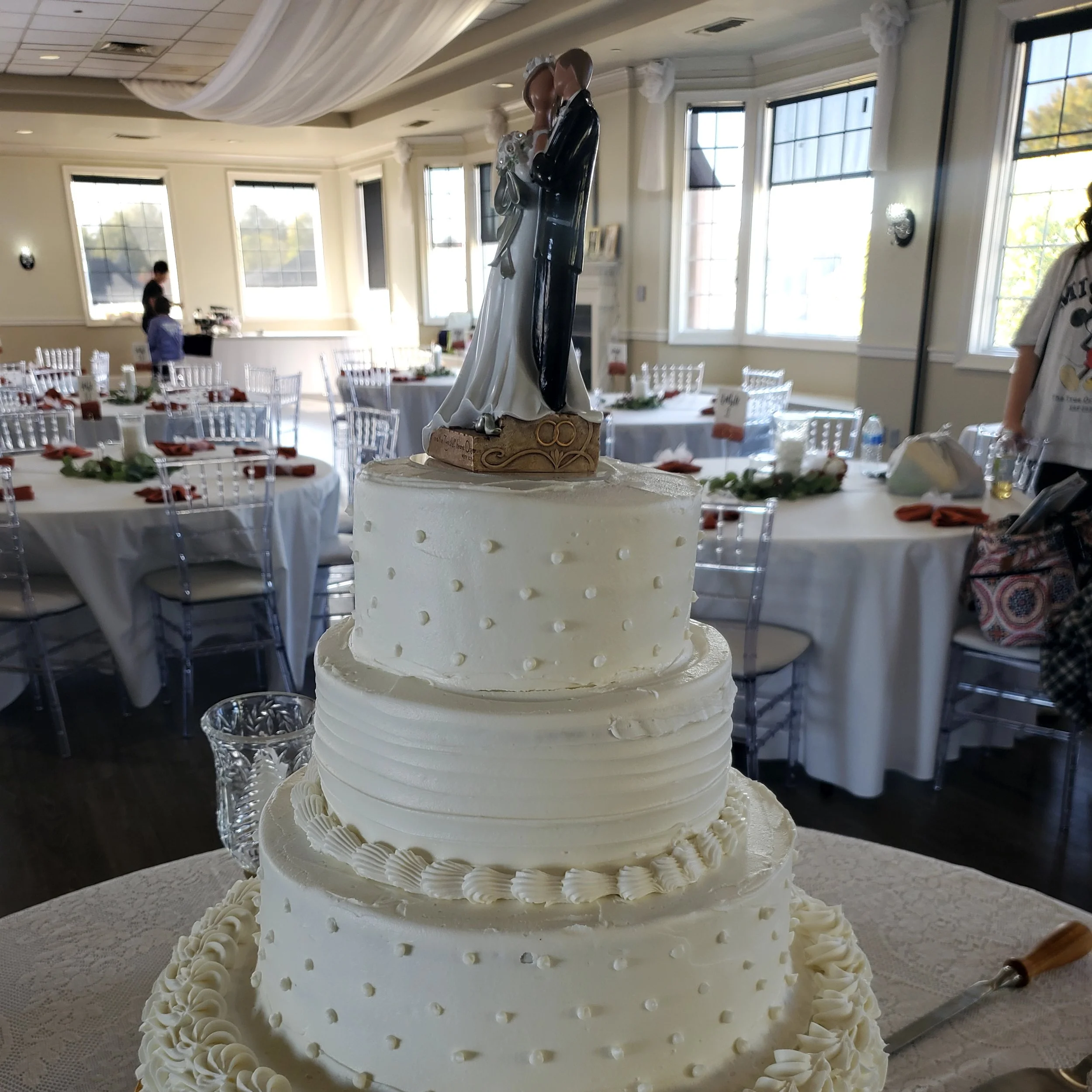 A wedding cake with four tiers topped with a bride and groom figurine cake topper, decorated with white icing and small decorative Lambeth scrolls.