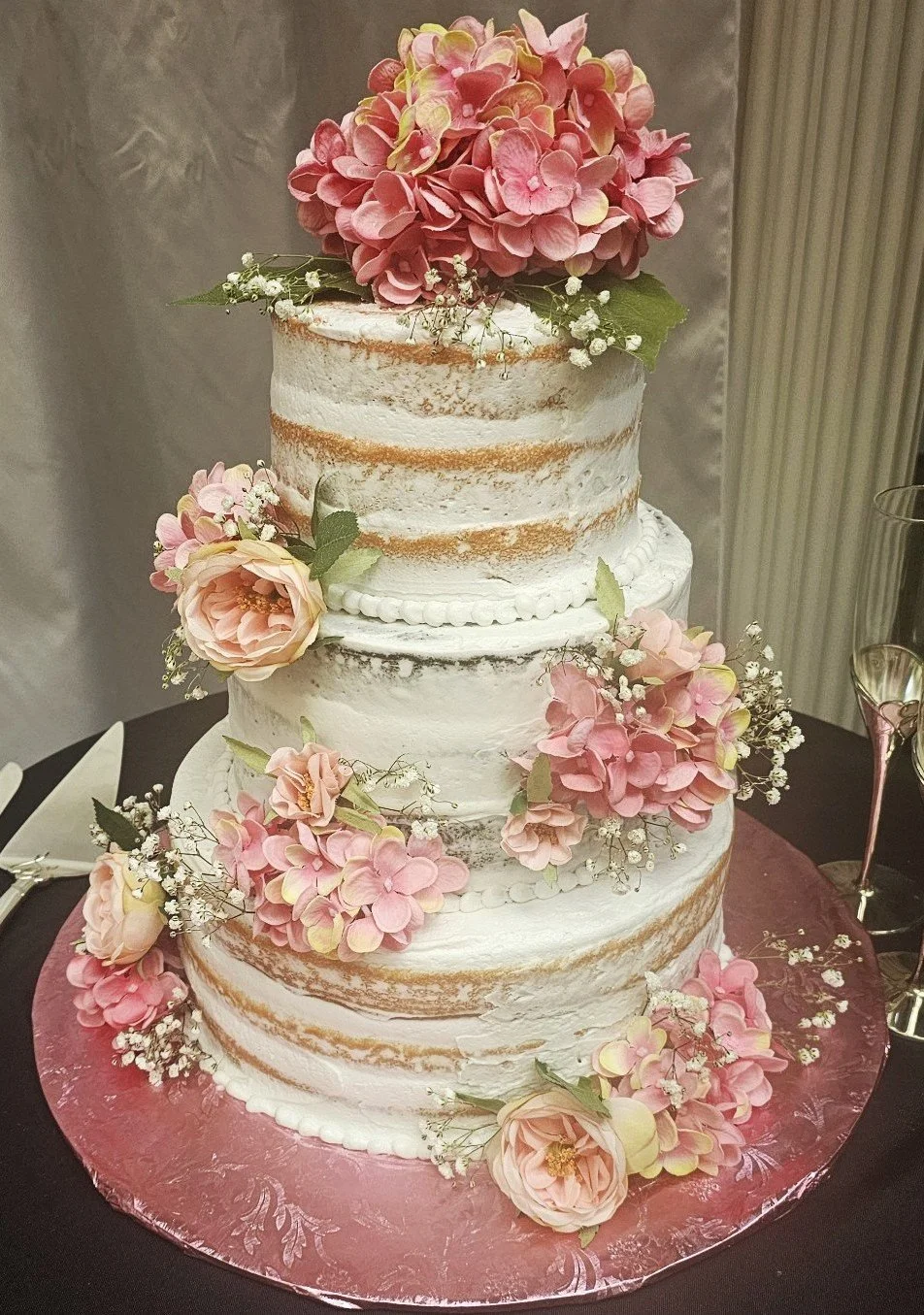 A three-tiered wedding cake with white crumb coat frosting and sparsely decorated with pink and cream hydrangea flowers, small white baby's breath, and peach roses, set on a pink decorative cake board.