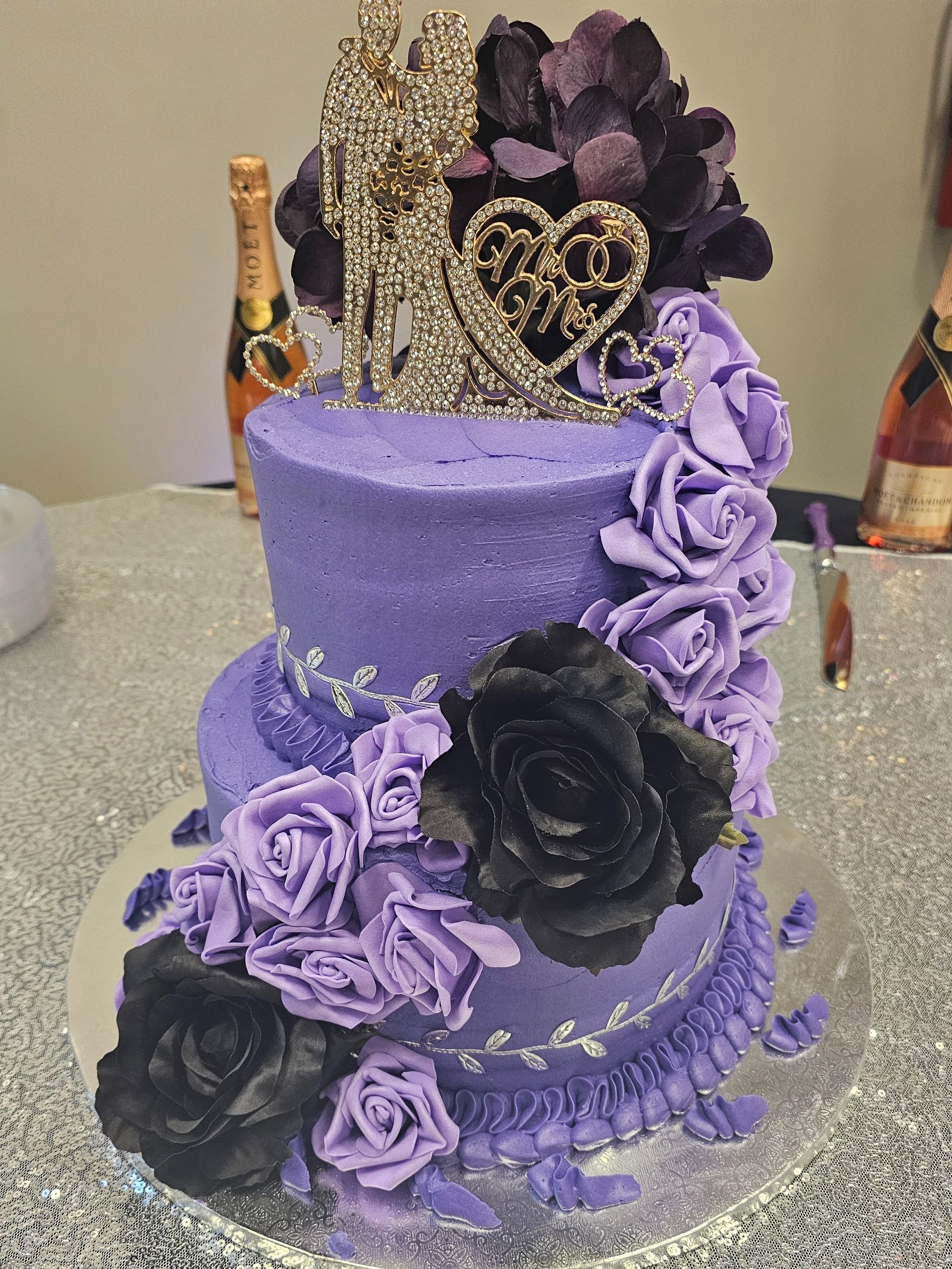 A three-tier purple wedding cake decorated with purple and black roses, silver leaf details, and a glittery 'Mr & Mrs' cake topper.
