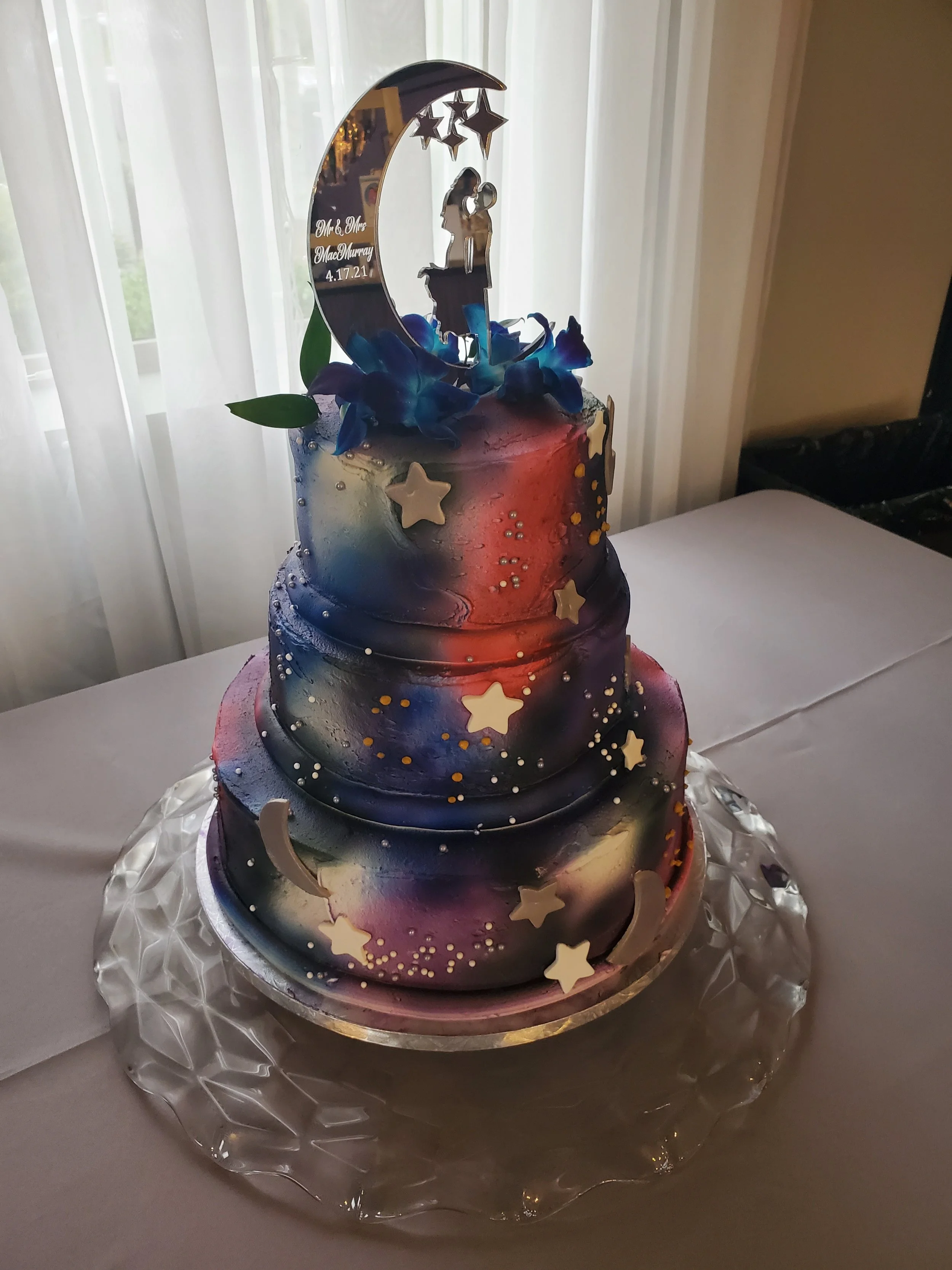 A three-tiered galaxy-themed birthday cake with blue, red, black, and purple swirled icing, decorated with star-shaped toppers, small decorative beads, and blue flowers. The cake topper features a silhouette of a girl sitting on a crescent moon with stars, and has personalization text with a date and names.
