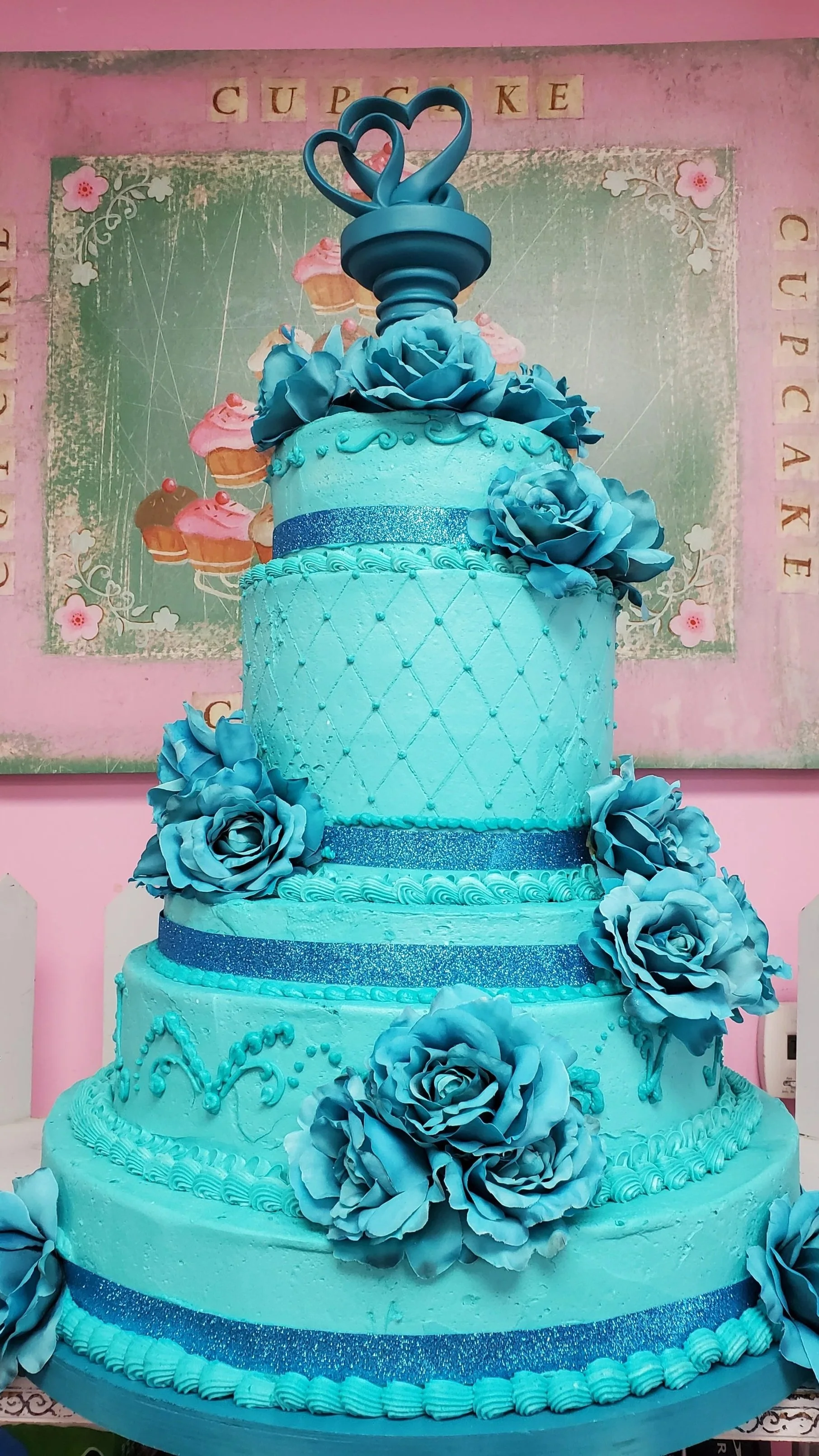 A four-tier monochrome teal cake wedding cake decorated with large blue roses and glittery blue ribbons, topped with a heart-shaped cake topper. The cake will serve 500.
