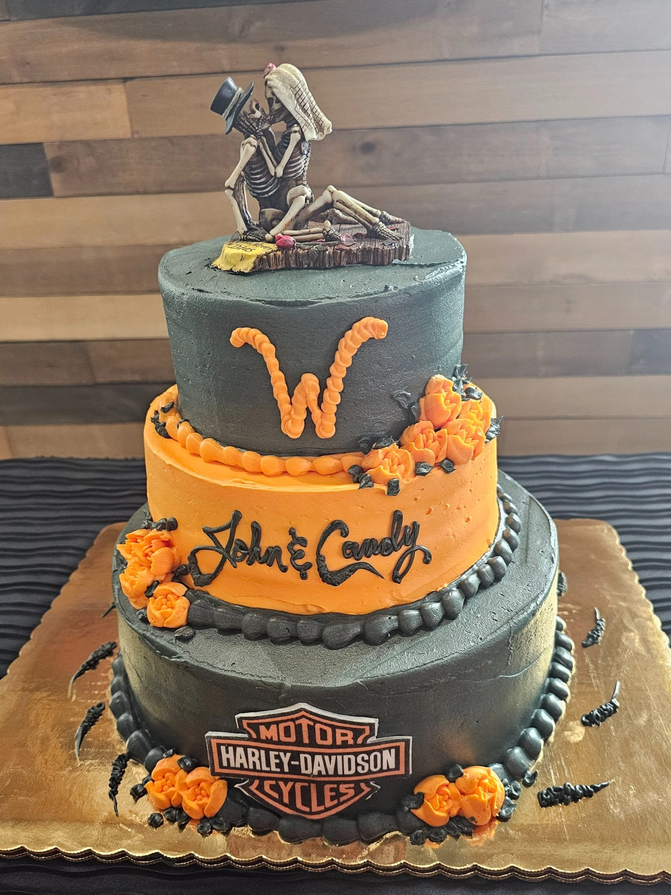 A three-tiered birthday cake with a skeleton couple topper, decorated in Harley-Davidson themes with black and orange colors, featuring the names John & Candy. Wild Wedding Cake!