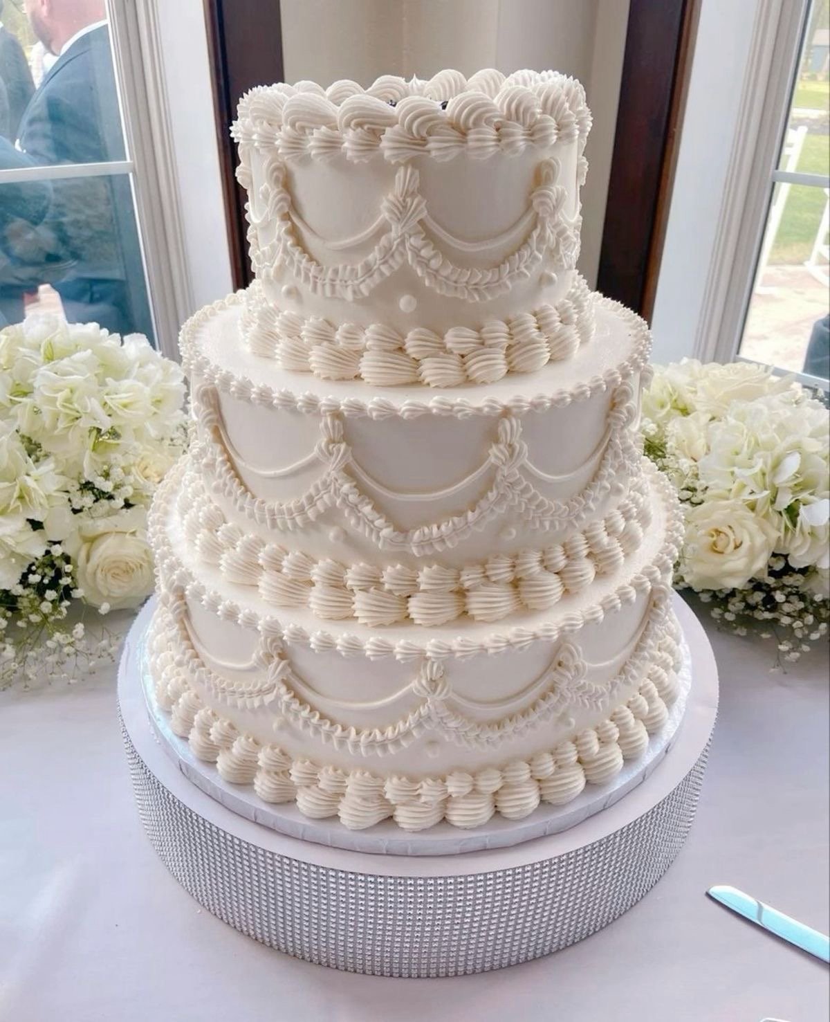 Three-tier white wedding cake with decorative Lambeth piping and floral accents, surrounded by white flowers.