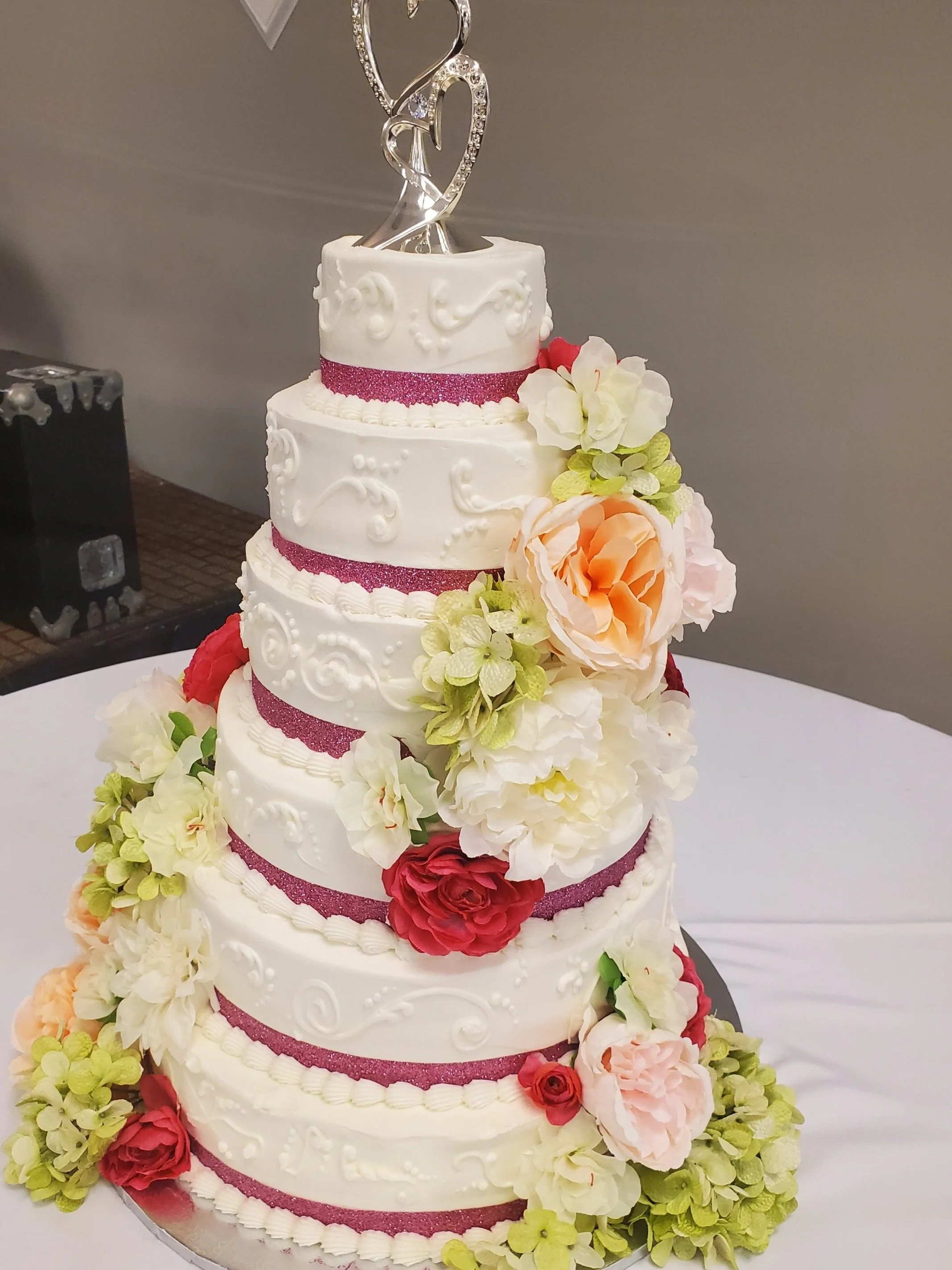 Tall, multi-tiered white wedding cake decorated with pink and white flowers and a silver heart-shaped cake topper.
