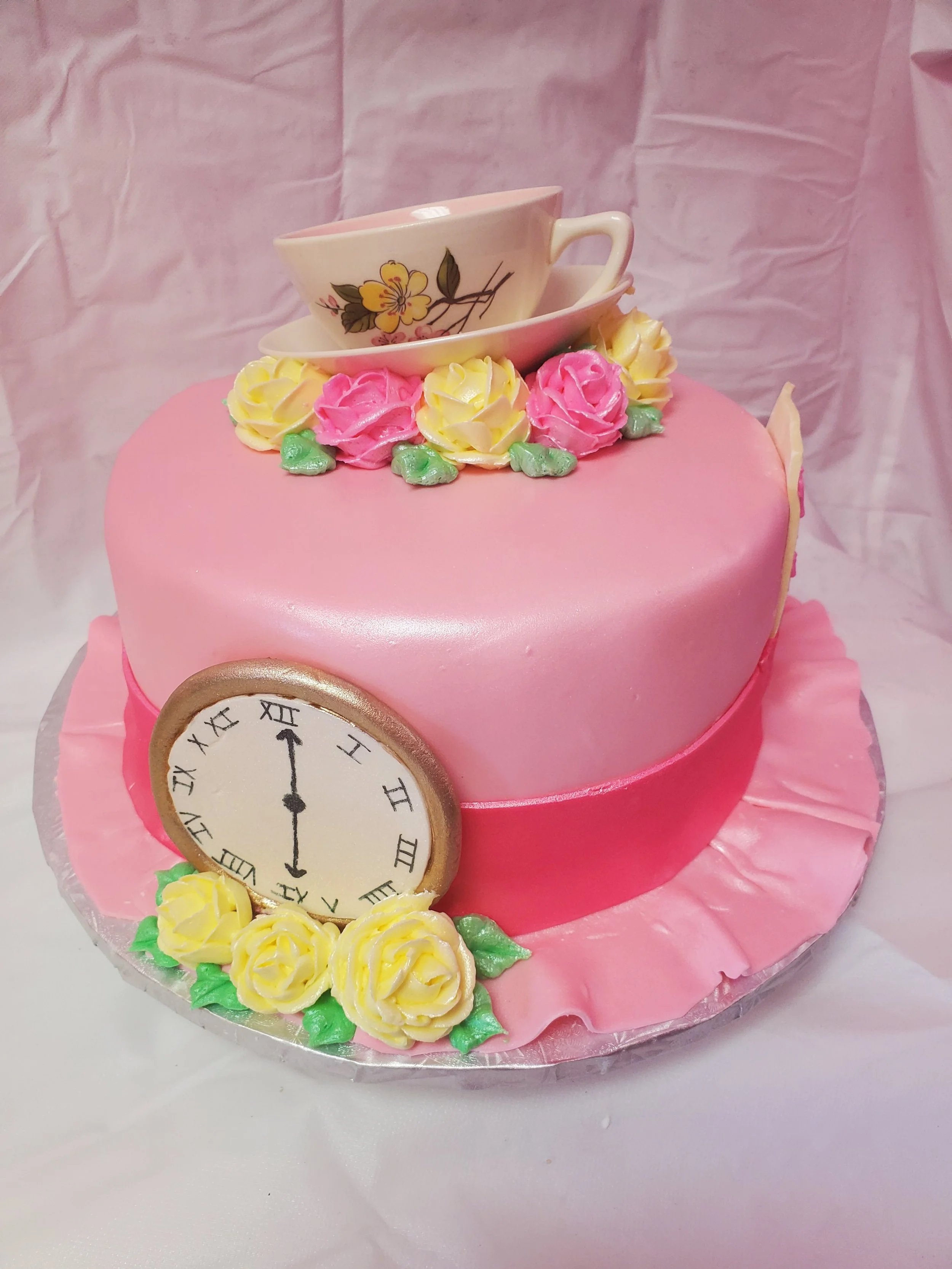 Pink fondant round cake decorated with yellow and pink frosting flowers, with actual china tea cup on top and a fondanr clock face on the front, set against a pink background.