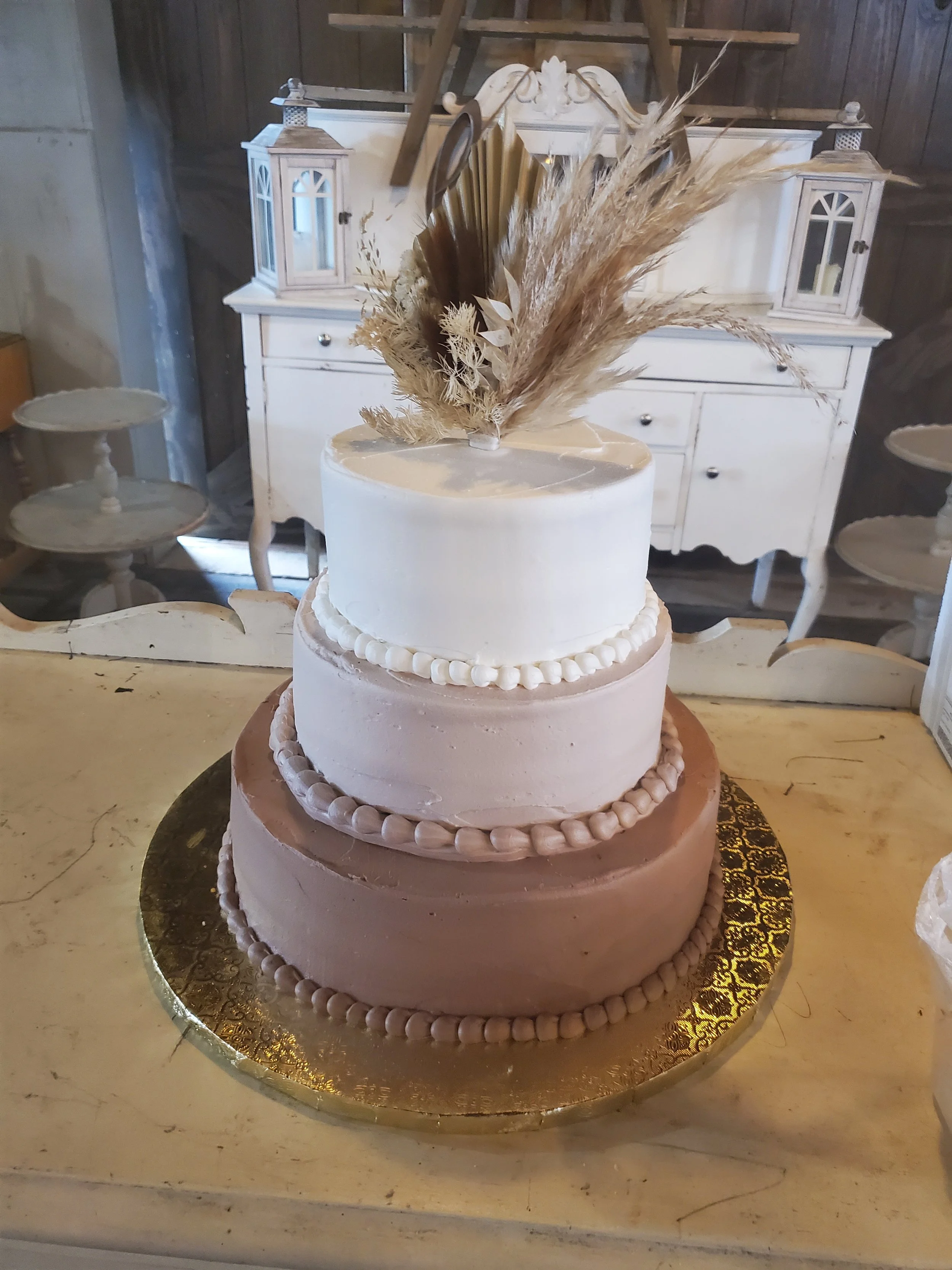 A three-tiered cake with white, pink, and peach frosting, decorated with piped borders and topped with dried pampas grass and other dried flowers, placed on a gold cake board.