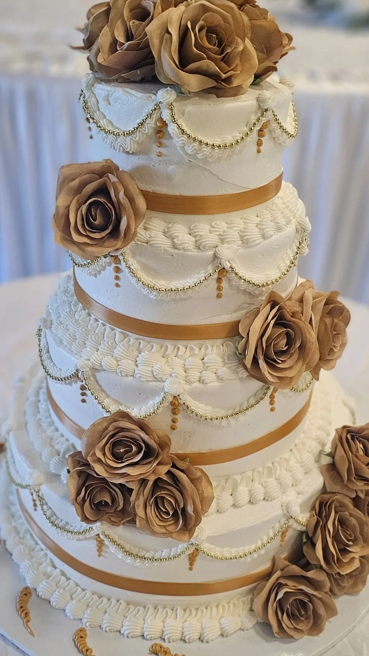 A tall, four-tiered wedding cake decorated with brown roses, gold beads, and cream-colored piping.