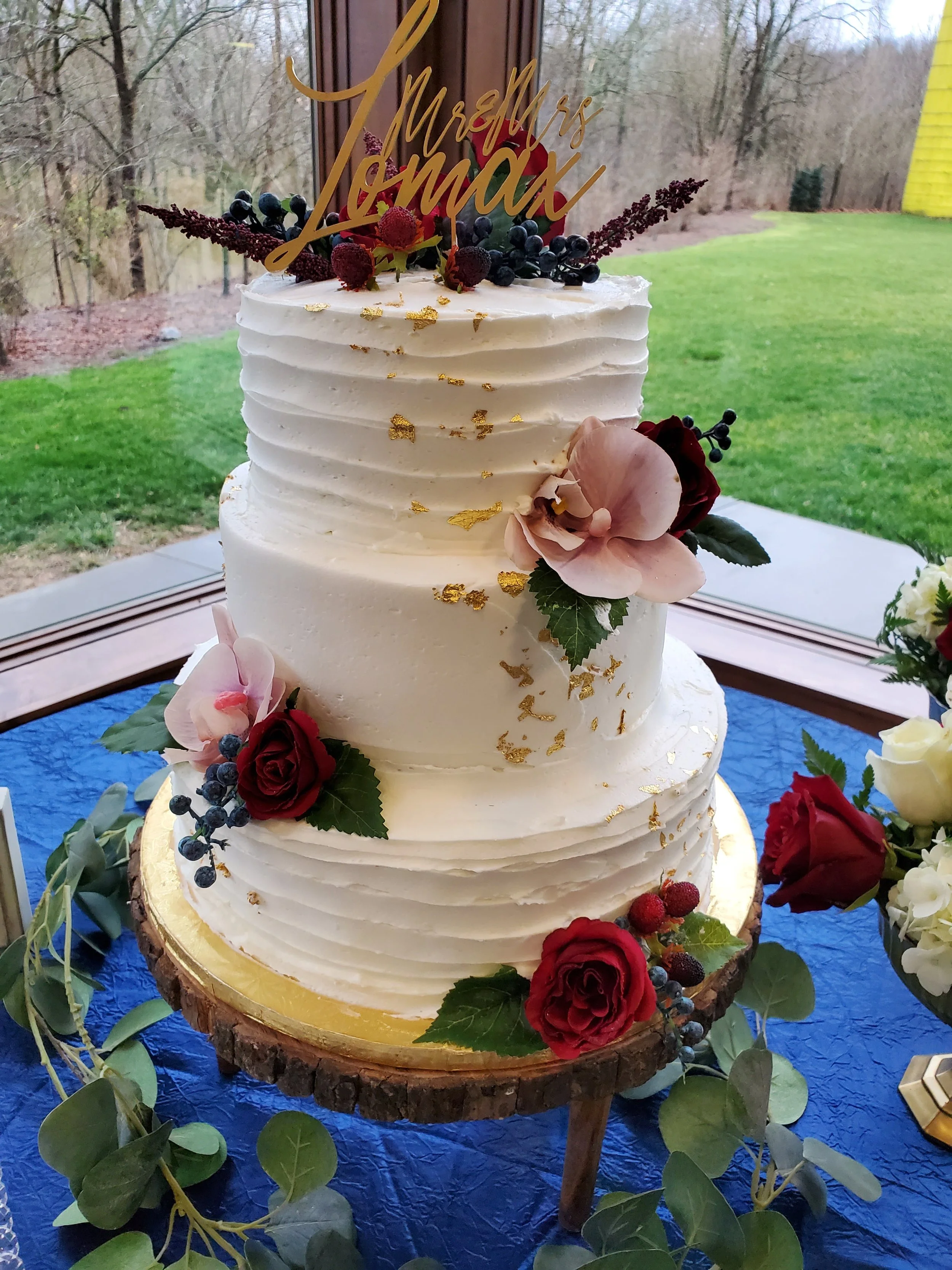 A three-tier white wedding cake decorated with flowers, berries, and gold leaf, topped with a gold 'Mrs. & Mr. Max' cake topper, displayed on a wooden slice stand.