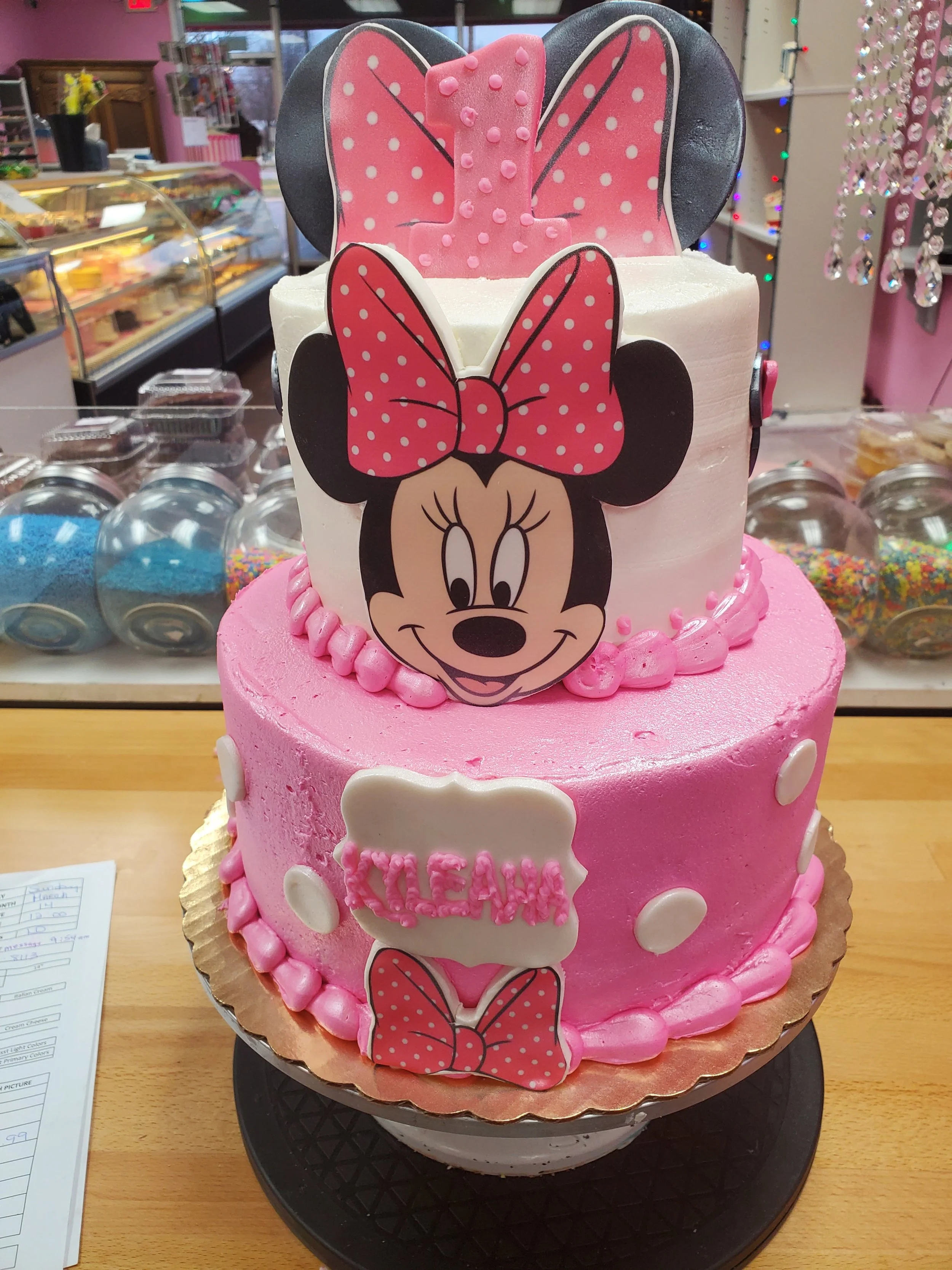 Three-tier pink birthday cake with M Mouse theme, featuring M Mouse faces with pink polka dot bows and white polka dots. The cake has decorations with the name 'KLEAH' in pink letters.