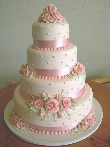 Four-tiered white wedding cake decorated with pink roses, green leaves, and pink satin ribbons beads, topped with a cluster of pink roses.