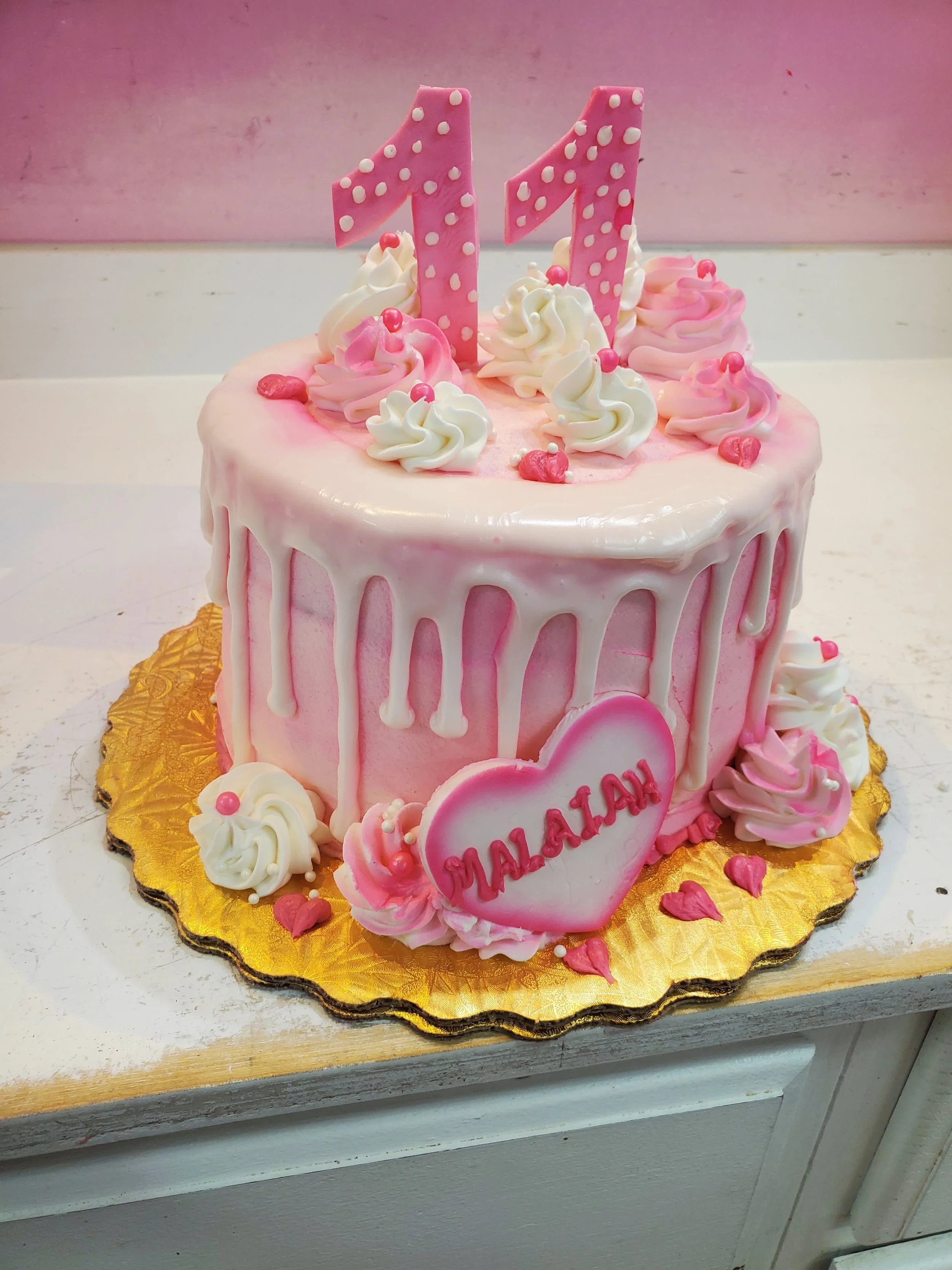 Pink birthday cake with white drip icing, pink and white swirls, and a pink heart with the name Malaiyah, topped with two pink number one candles.